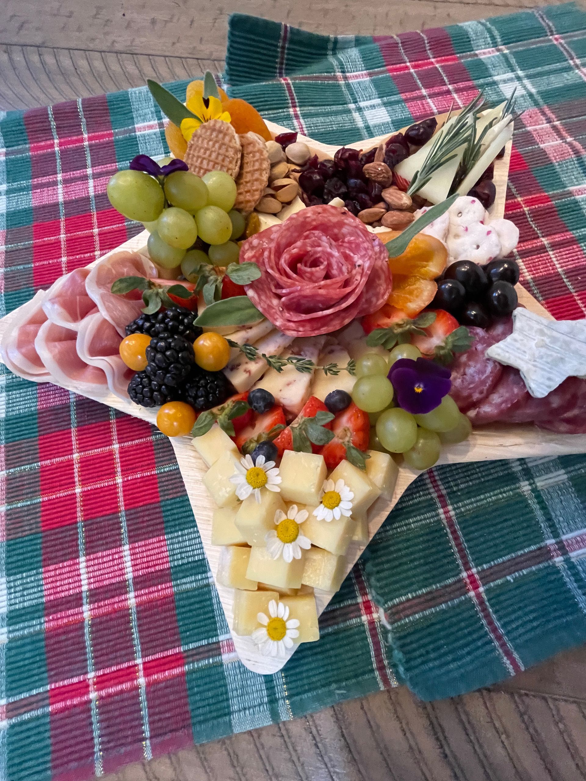 Star-shaped charcuterie board: cheeses, meats, fruits, nuts, and flowers on plaid cloth.
