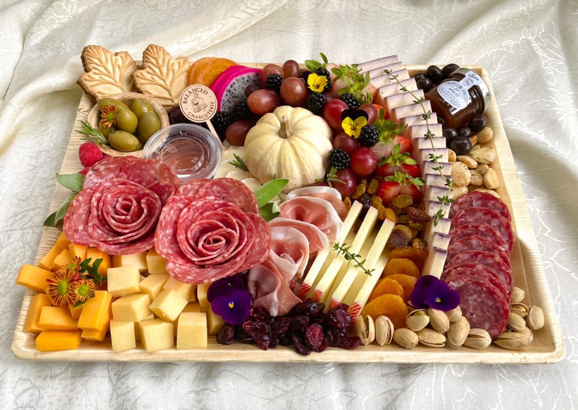 Charcuterie board with salami roses, cheeses, fruits, nuts, and a small pumpkin, arranged on a cream-colored platter.
