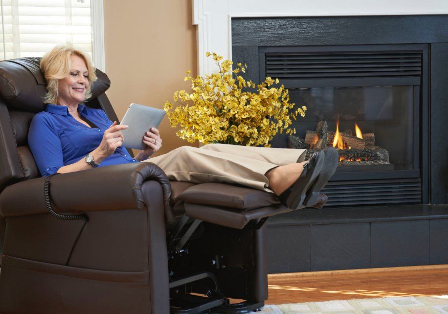 A woman is sitting in a recliner using a tablet in front of a fireplace.