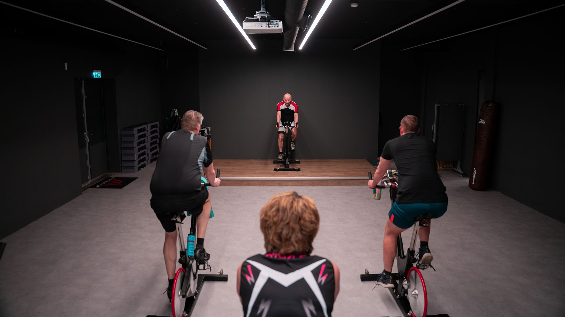 Vier mensen op hometrainers in een studio, tegenover een instructeur op een podium. Donkere ruimte, plafondverlichting.
