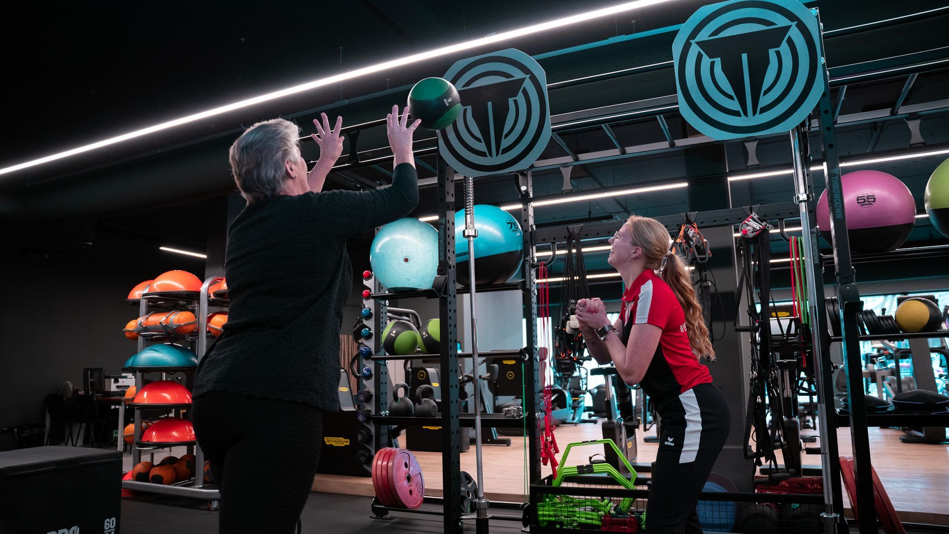 Twee mensen gooien in een gymzaal een medicijnbal. De persoon links gooit de bal omhoog naar de persoon rechts.