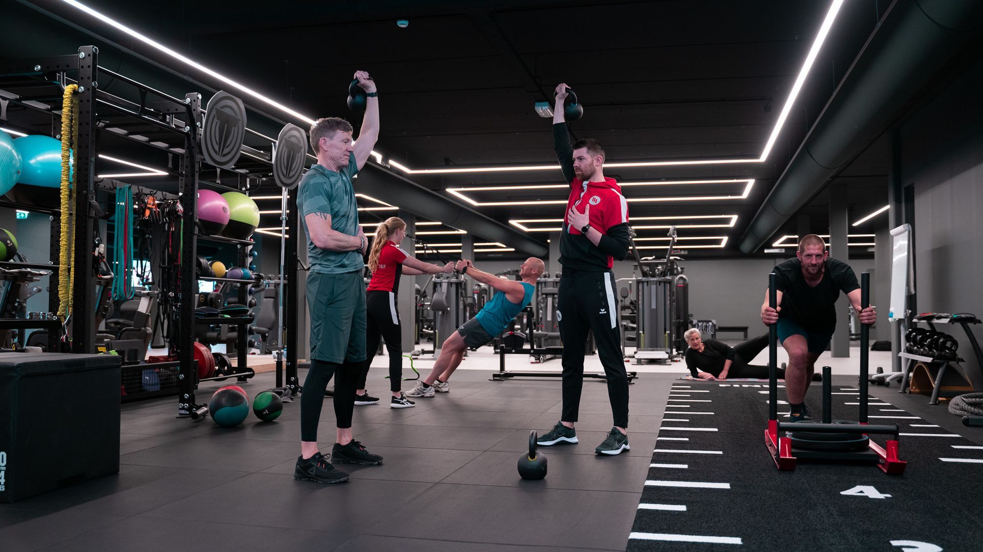 Mensen trainen met kettlebells in een sportschool onder felle stripverlichting.