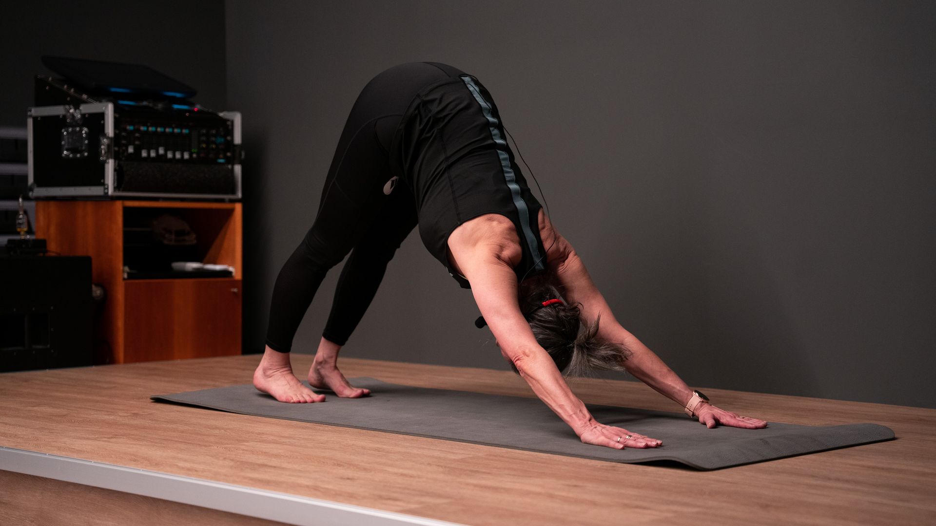 Persoon in de neerwaartse hondhouding op een yogamat. Zwarte legging en top, grijze mat, donkere achtergrond.