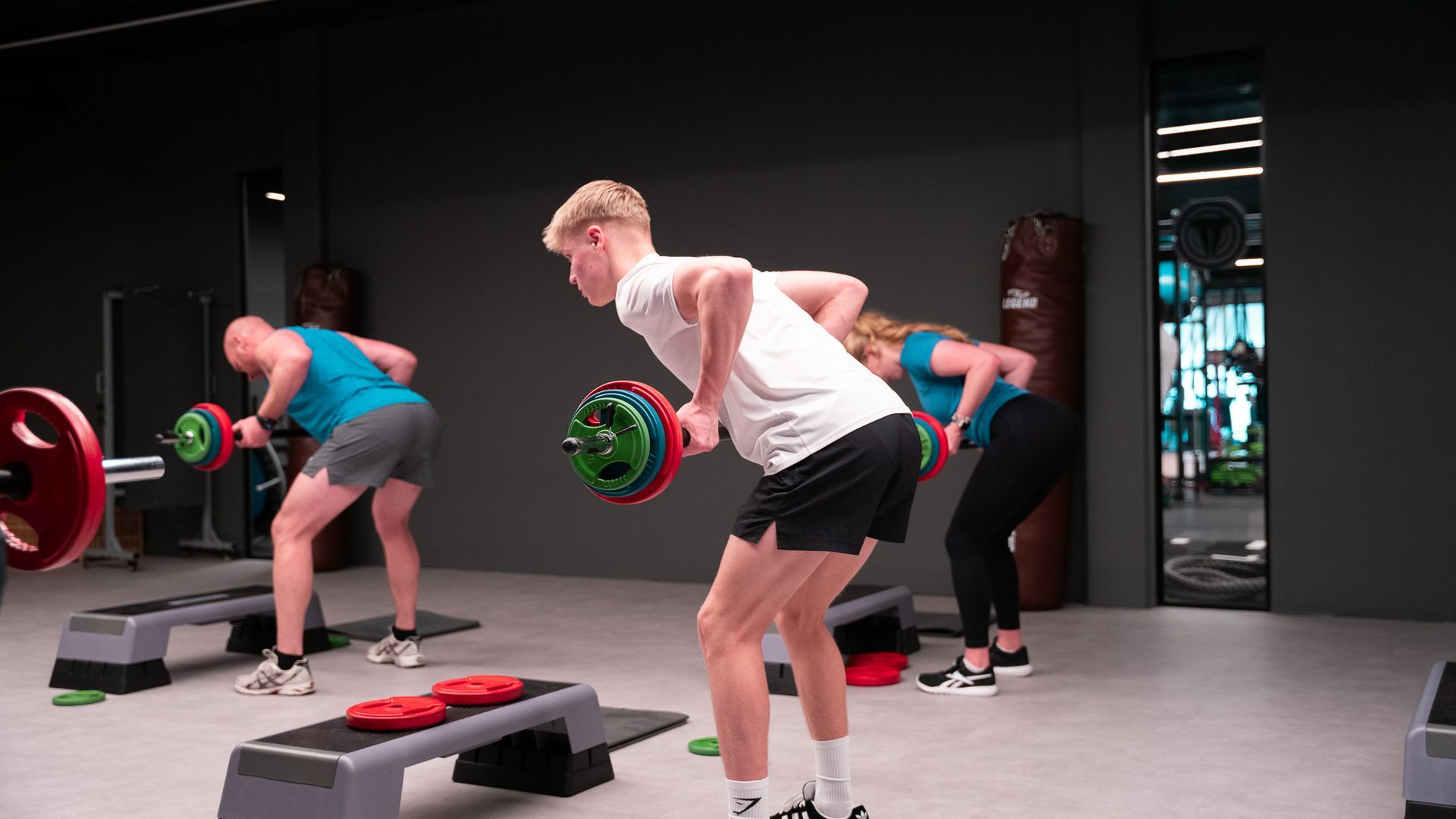 Drie mensen doen barbell rows in een sportschool, waarbij ze gewichtsschijven op een verhoging gebruiken.