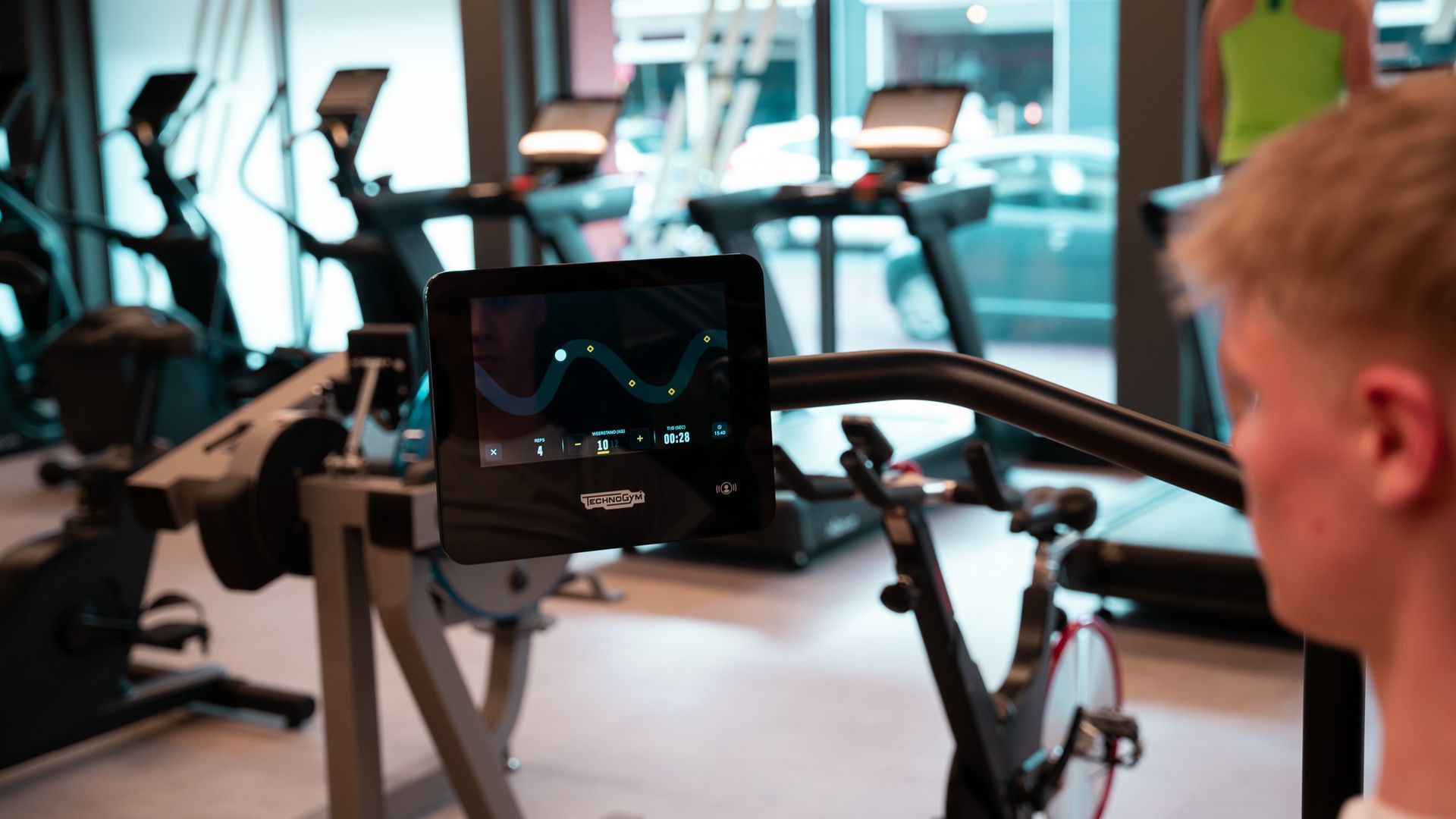 Een man gebruikt een hometrainer en kijkt naar een scherm in een sportschool waarop trainingsgegevens worden weergegeven.