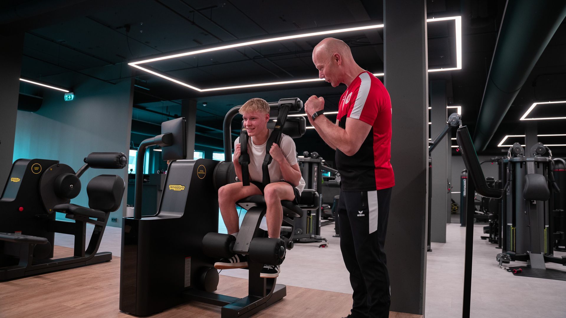 Een man helpt iemand bij het gebruik van een beenstrekapparaat in een sportschool. Beiden glimlachen.
