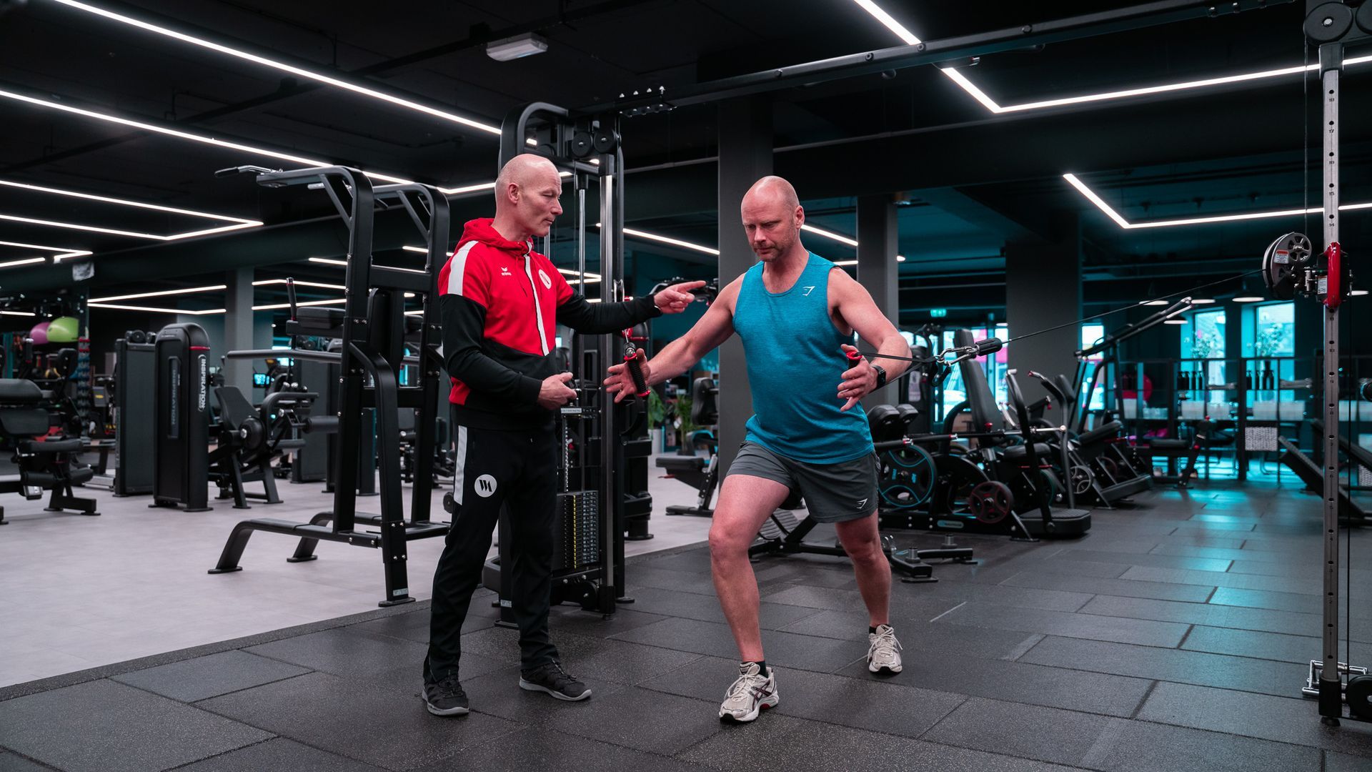 Een man doet oefeningen in de sportschool met een trainer; de vloer is zwart, gewichten zijn op de achtergrond te zien.