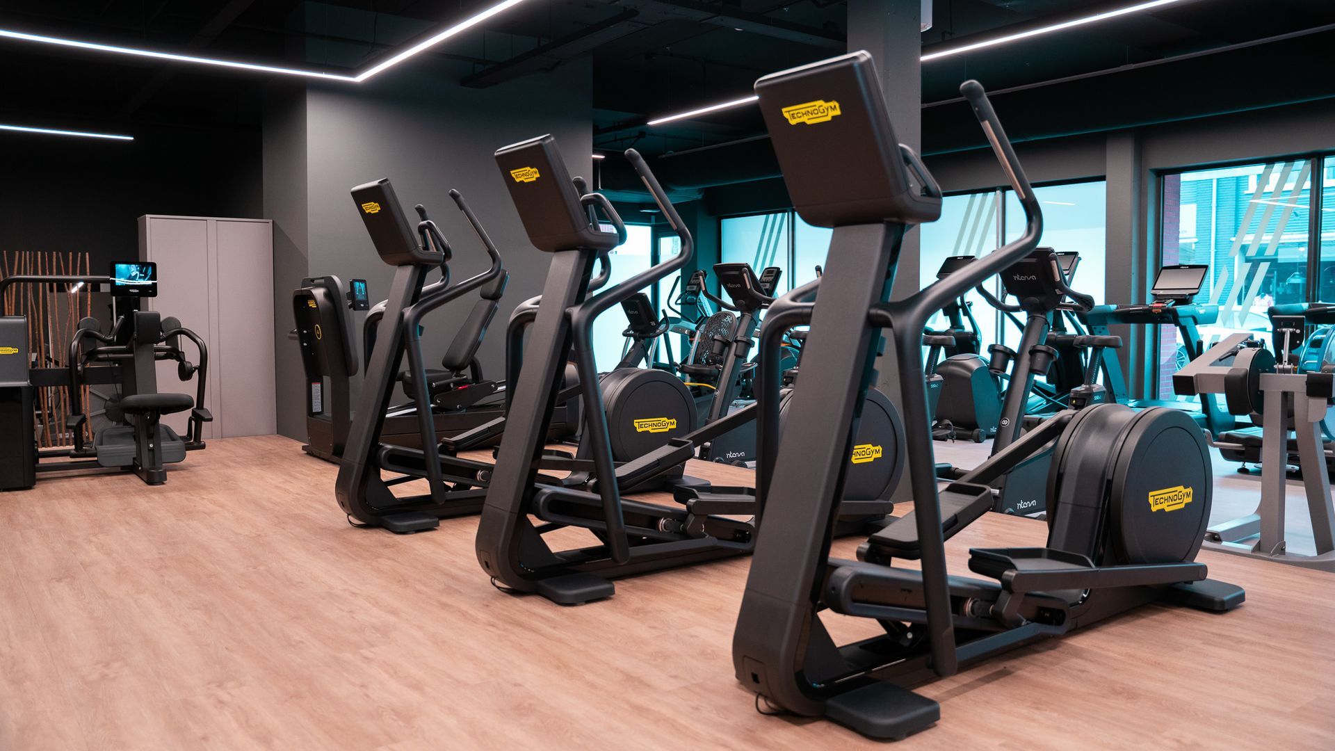 Een rij zwarte crosstrainers in een sportschool met houten vloer en donkere muren.
