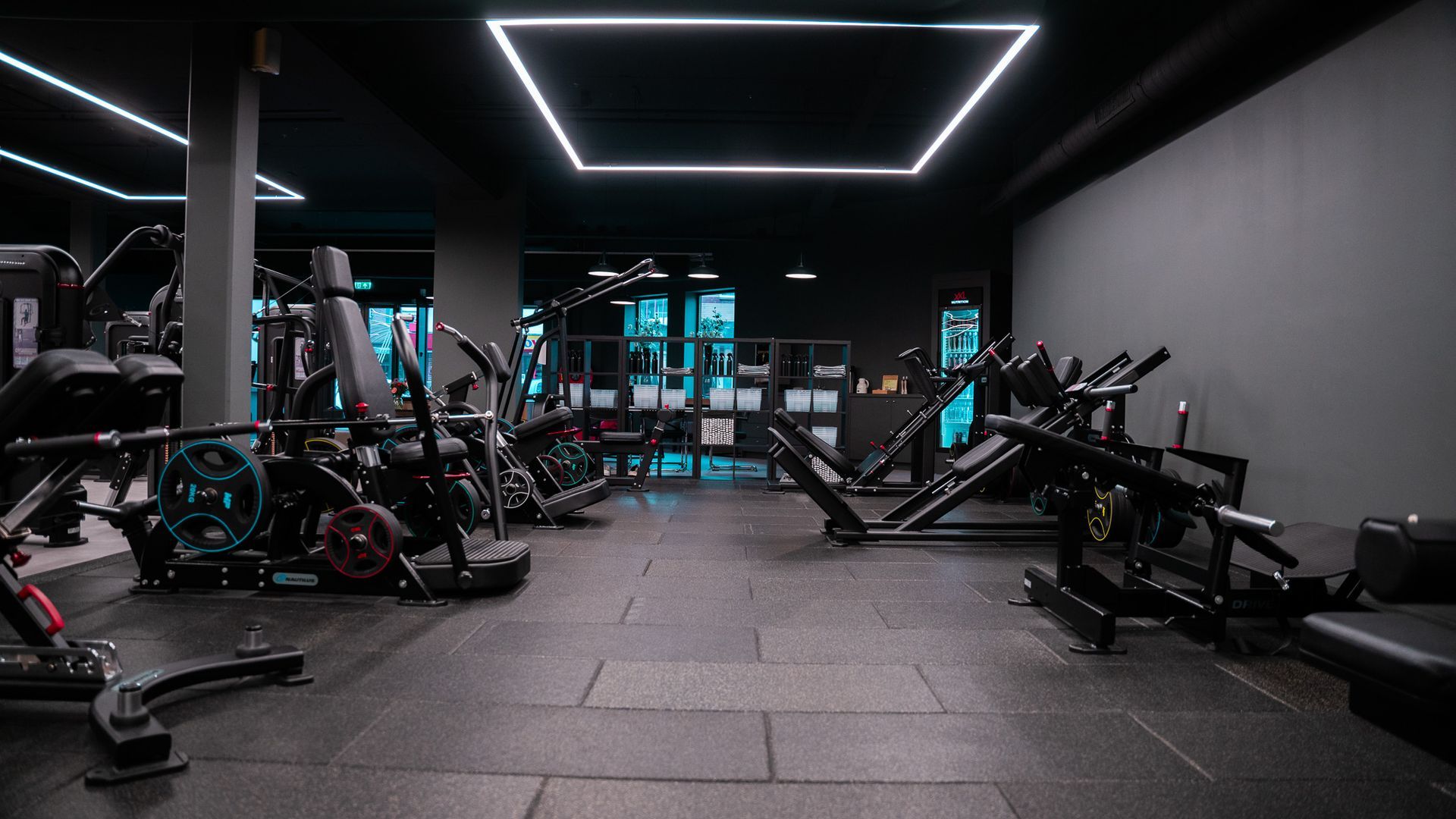 Interieur van een sportschool met diverse fitnessapparaten, grijze muren en felle plafondlampen.