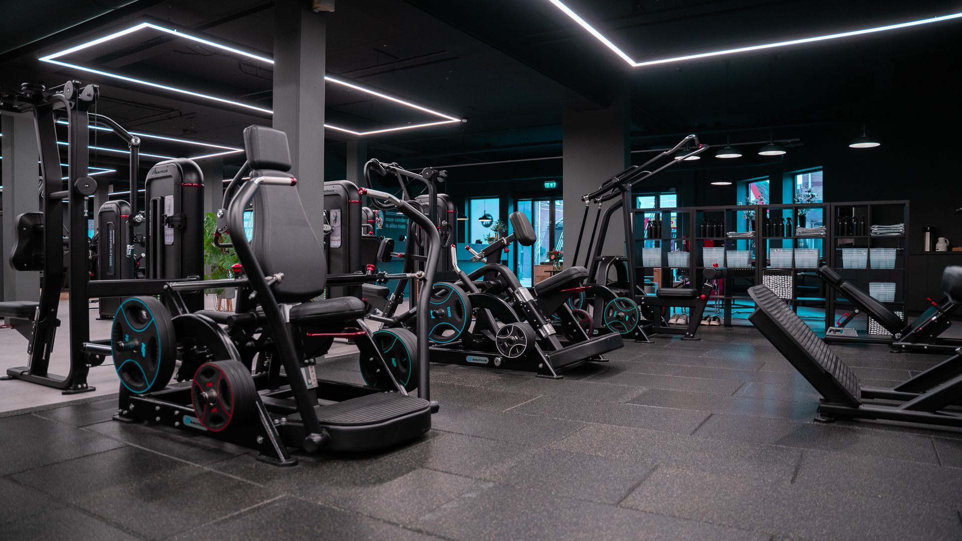 Sportschool met diverse fitnessapparaten, een donker interieur, tl-verlichting en zwarte apparatuur.