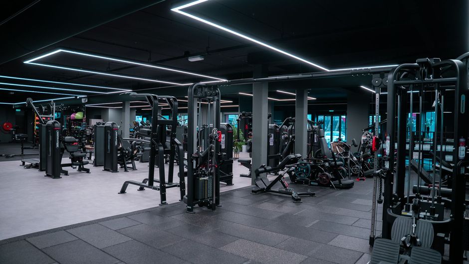 Interieur van een sportschool met diverse gewichtsapparaten en -uitrusting, onder een zwart plafond met neonverlichting.