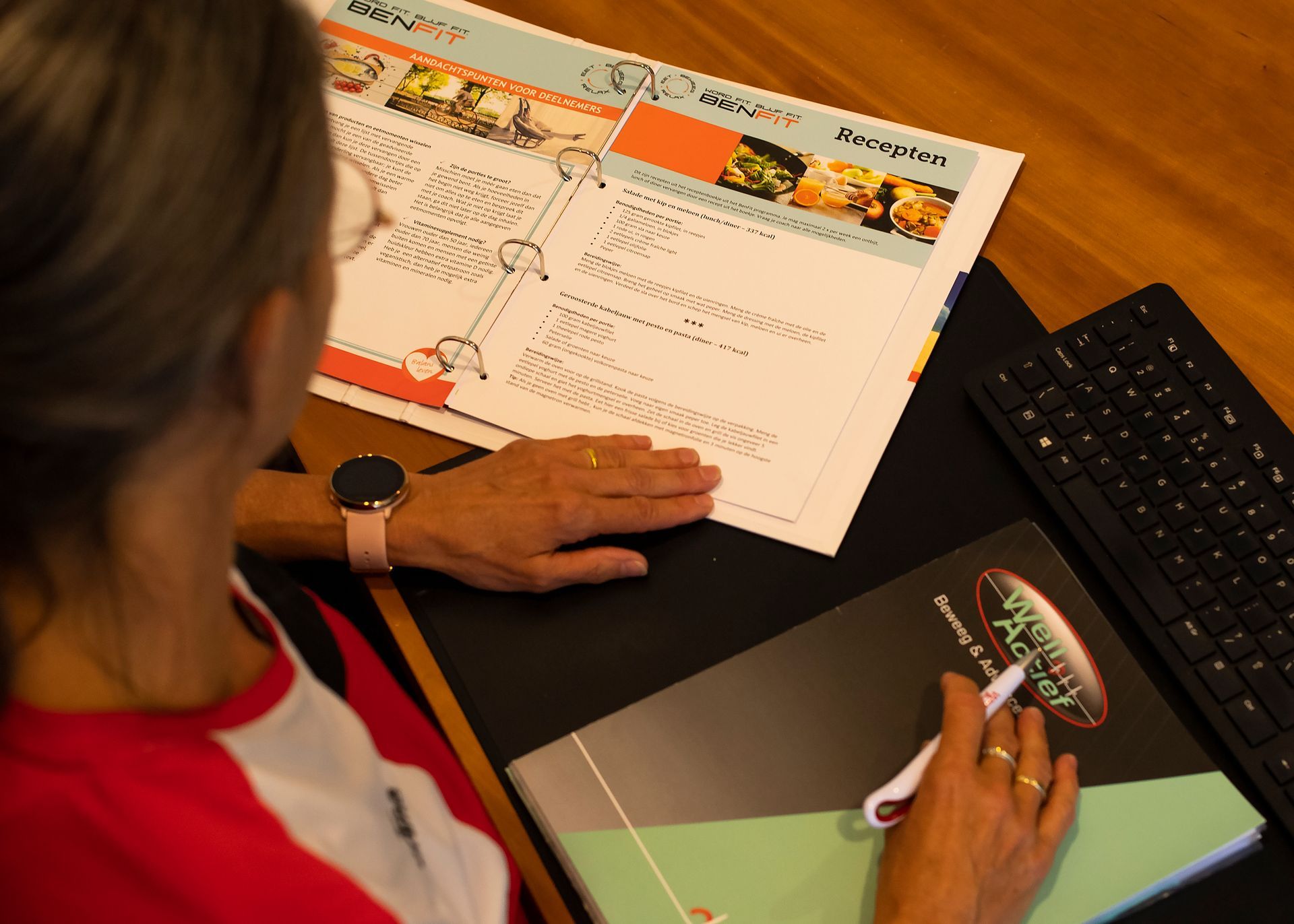 Een persoon met een bril en een smartwatch kijkt naar papieren op een bureau met een toetsenbord en een map.
