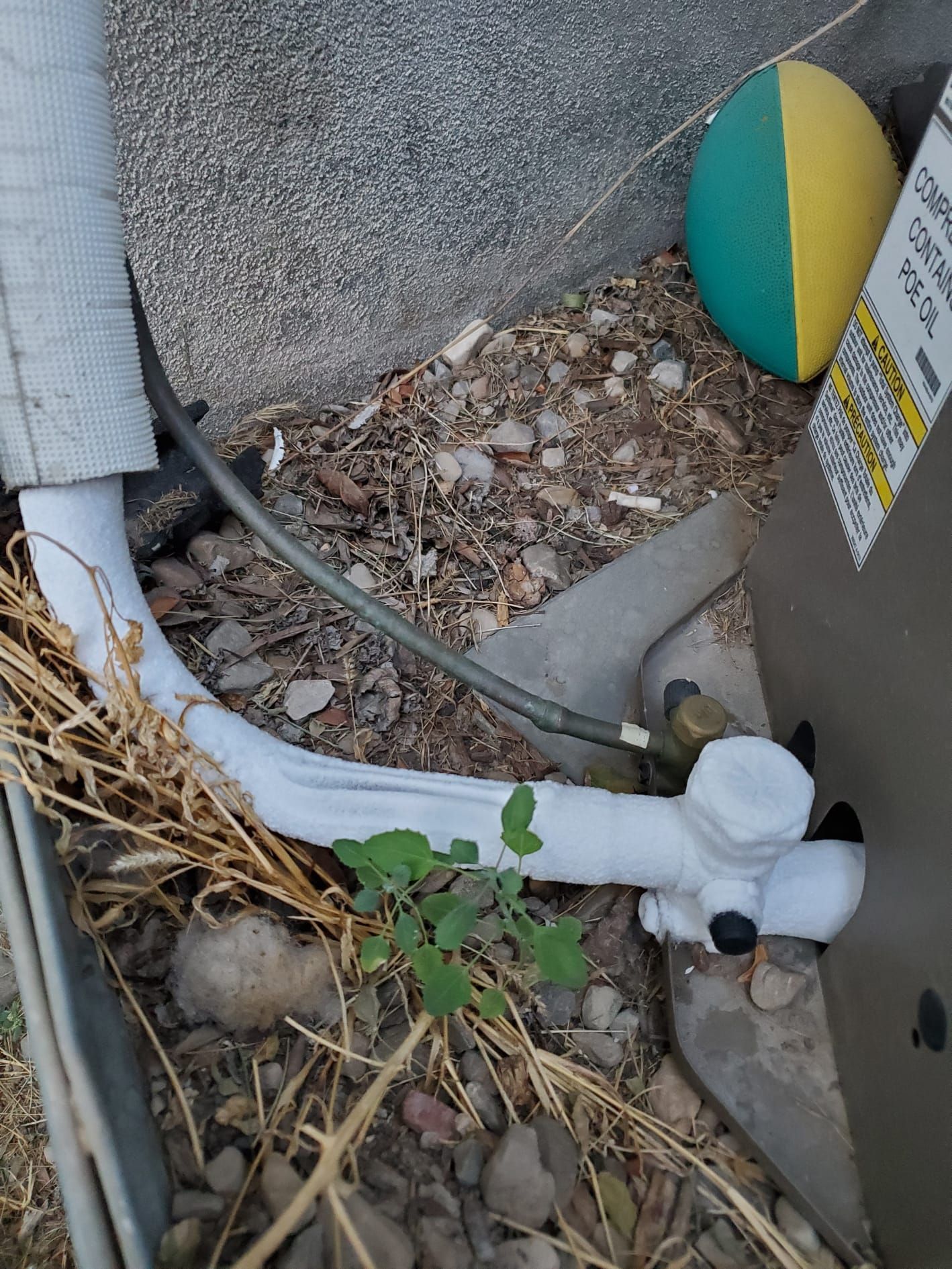 Close-up of white and black pipes and a cable in gravel beside a concrete wall, with dry leaves and weeds.
