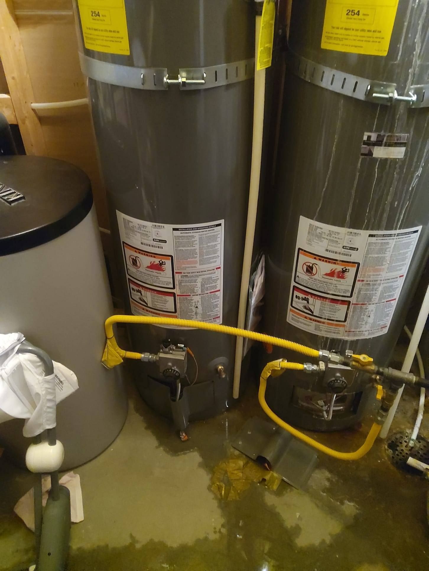 Two gray water heaters in a utility closet with yellow gas lines and a small amount of water on the floor