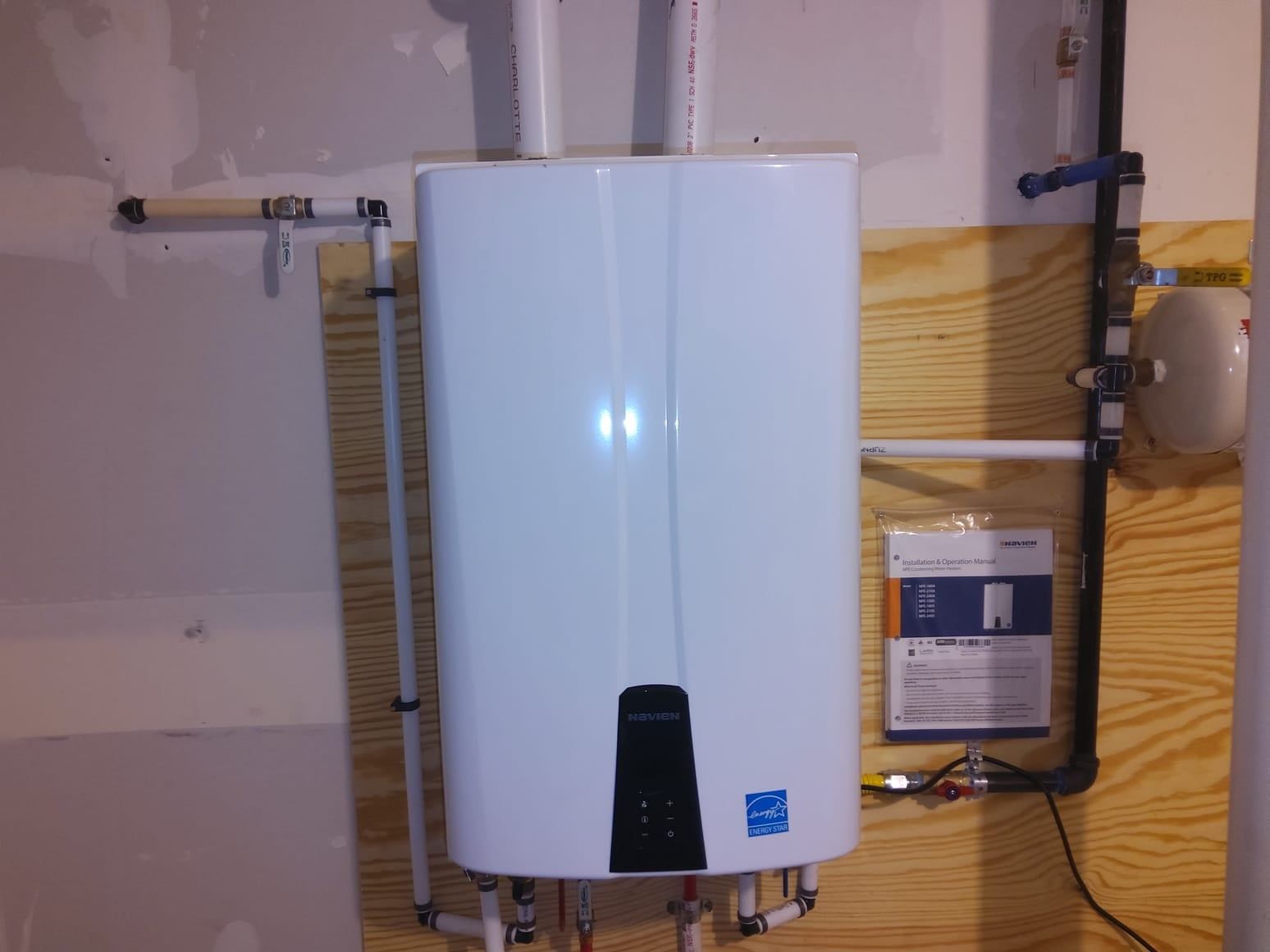 White wall-mounted tankless water heater with pipes in a utility room, next to insulation and wiring.