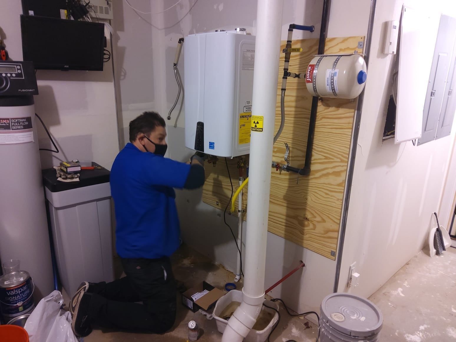 Technician kneeling beside a wall-mounted water heater, working on pipes in a utility room