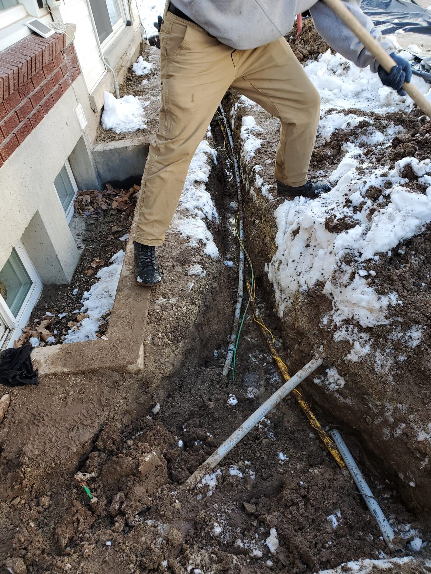 Worker standing in a muddy trench beside a house, with snow and exposed pipes around him.