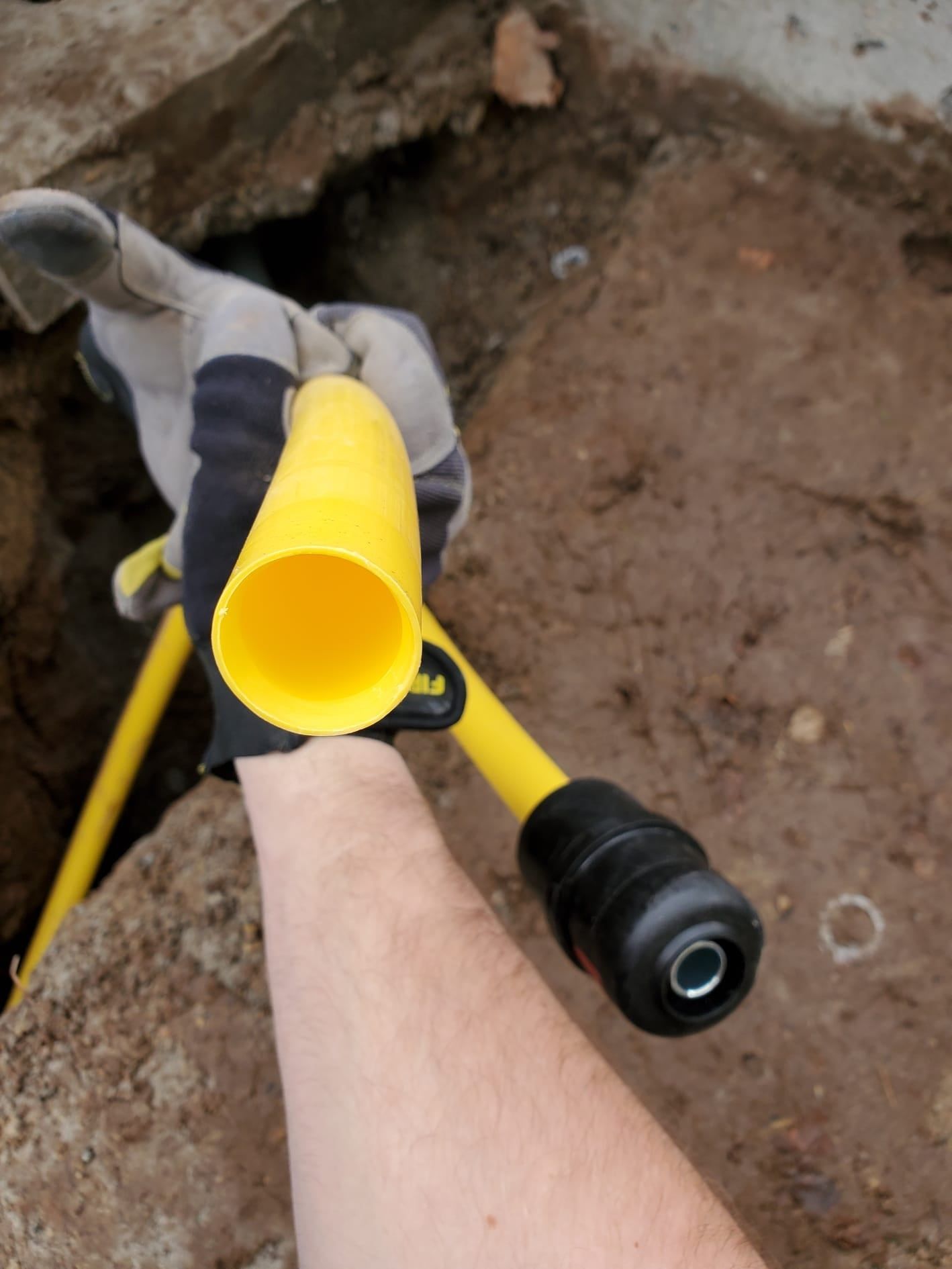 Gloved hand holding yellow and black pipe or cable over a dirt trench outdoors