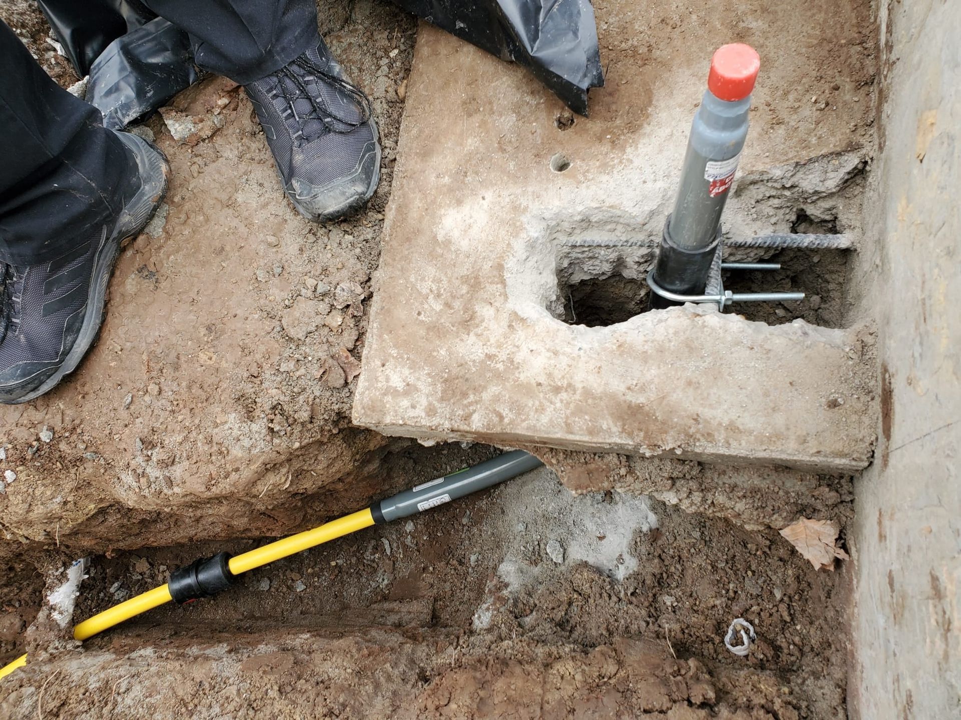 A hand tool beside a concrete footing with a metal anchor bolt and orange cap in a dirt excavation