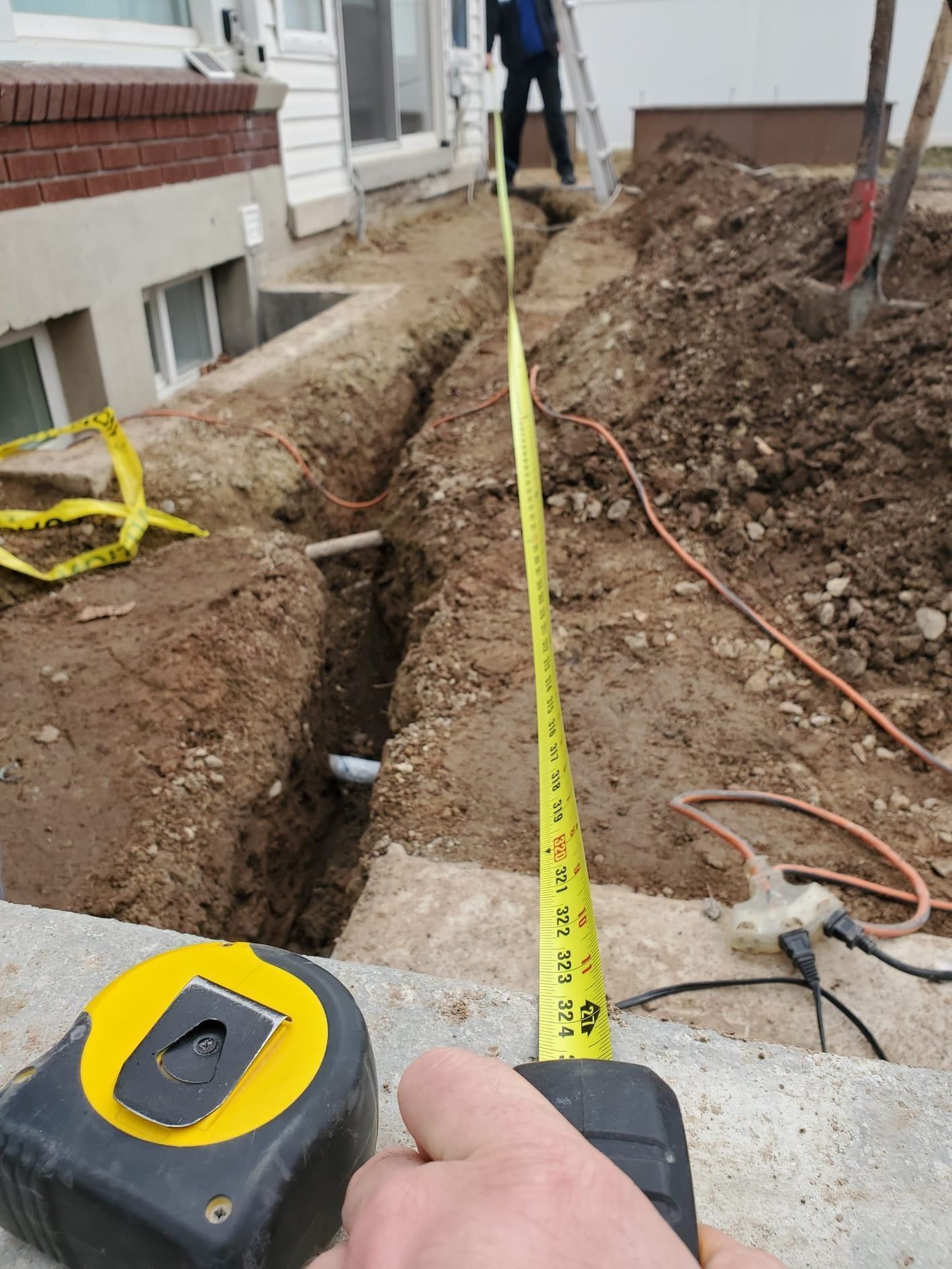 Measuring tape stretched over a trench beside a building, with dirt piles and a hose on the ground