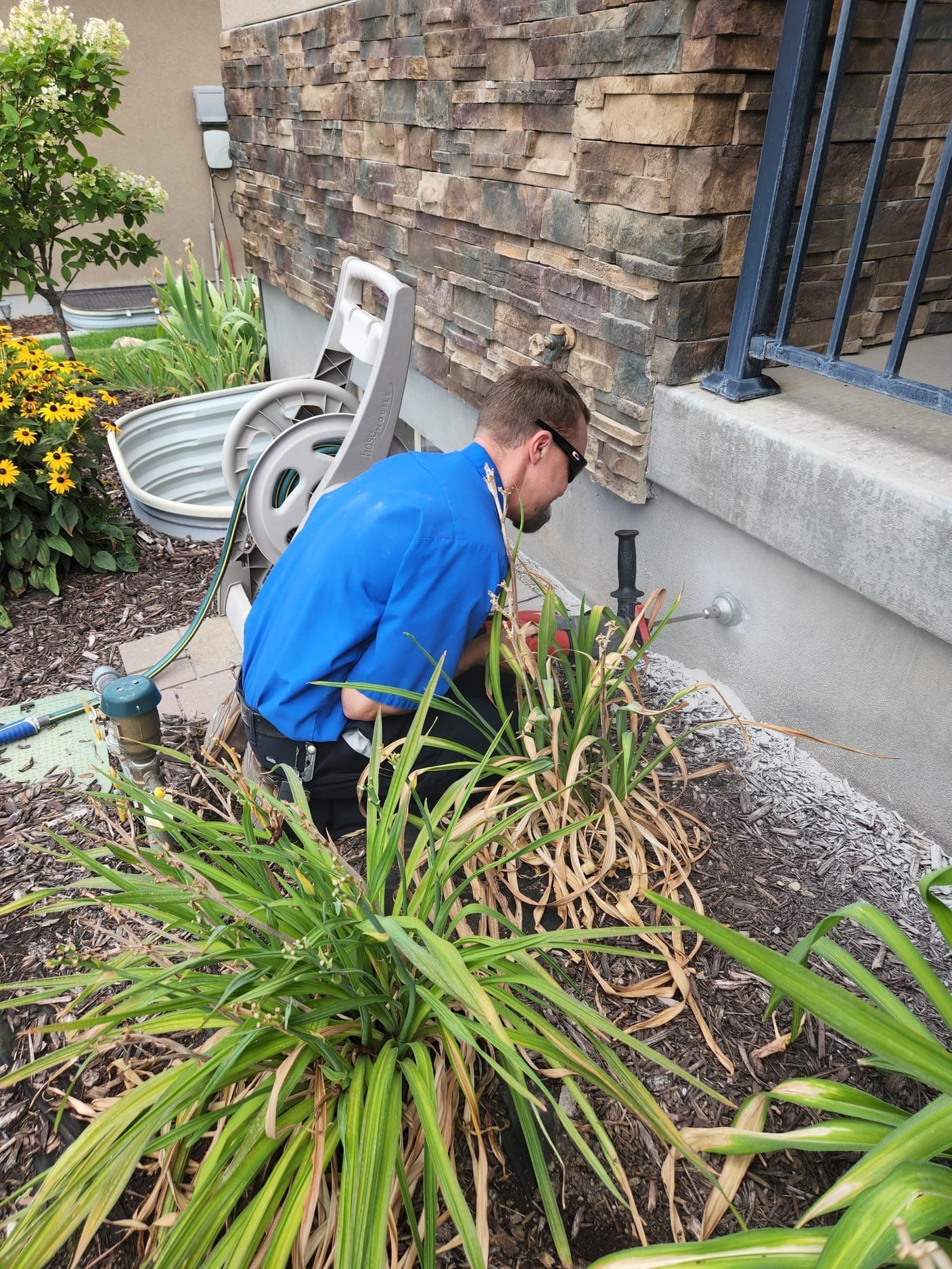 Person crouching beside a house foundation, weeding a flower bed near a stone wall and bench.