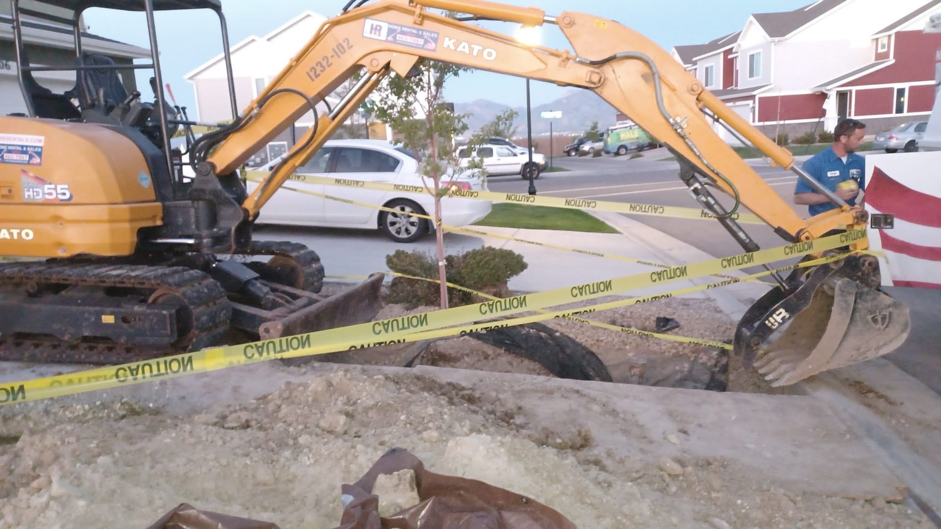 Yellow excavator at a residential street construction site, digging near caution tape and large rocks.