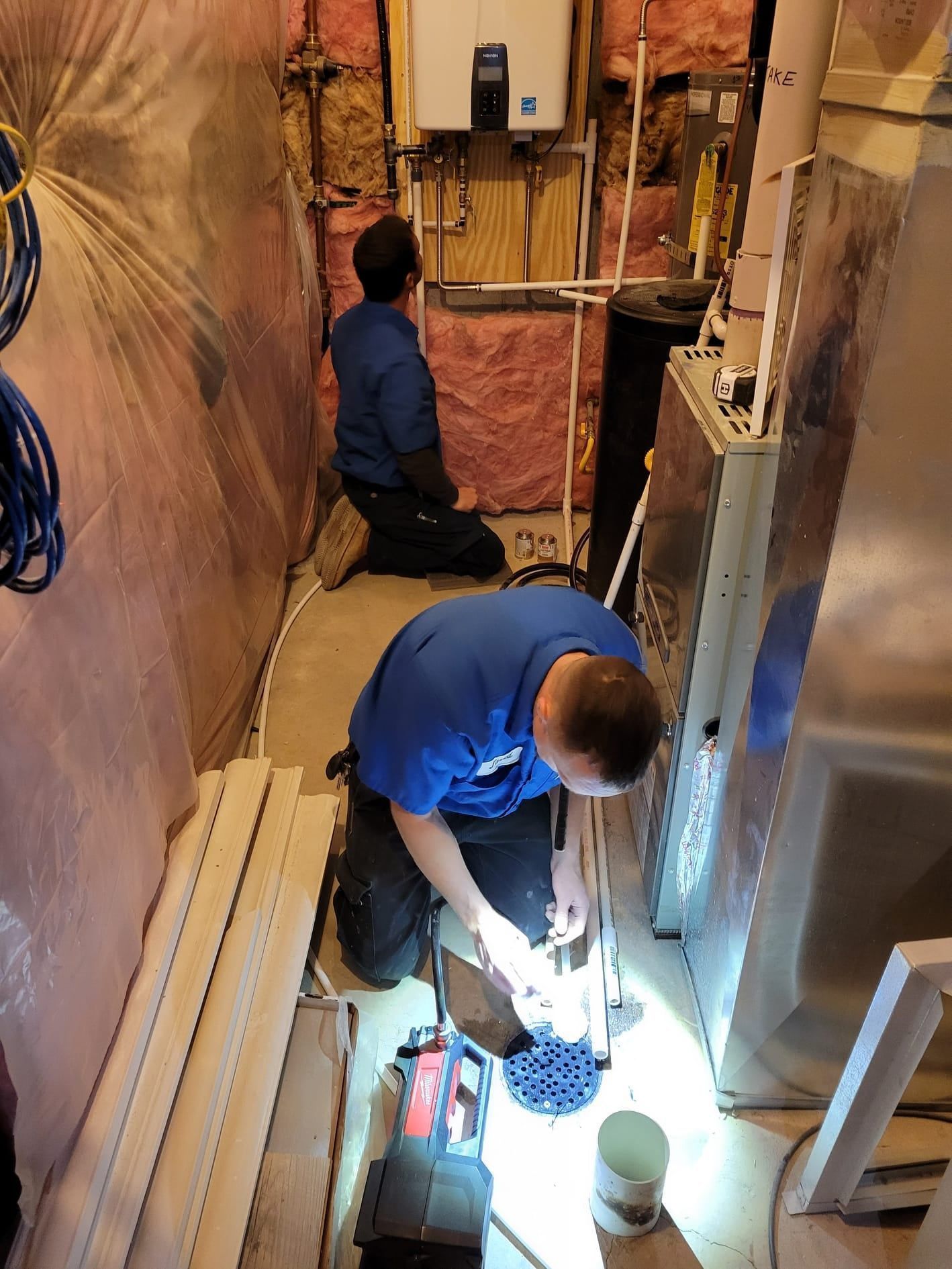 Two workers in a narrow utility space inspecting exposed pipes and wiring with a flashlight.
