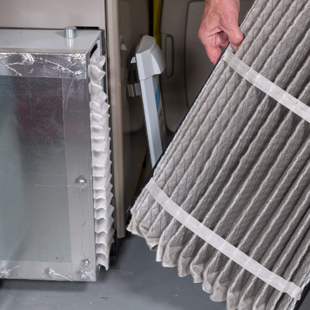 Hand holding a pleated HVAC air filter beside a metal duct and another filter unit