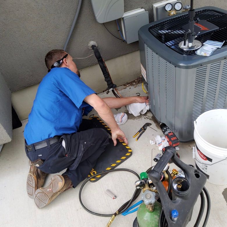 Technician kneels beside HVAC unit, repairing components with tools and gauges on the ground.