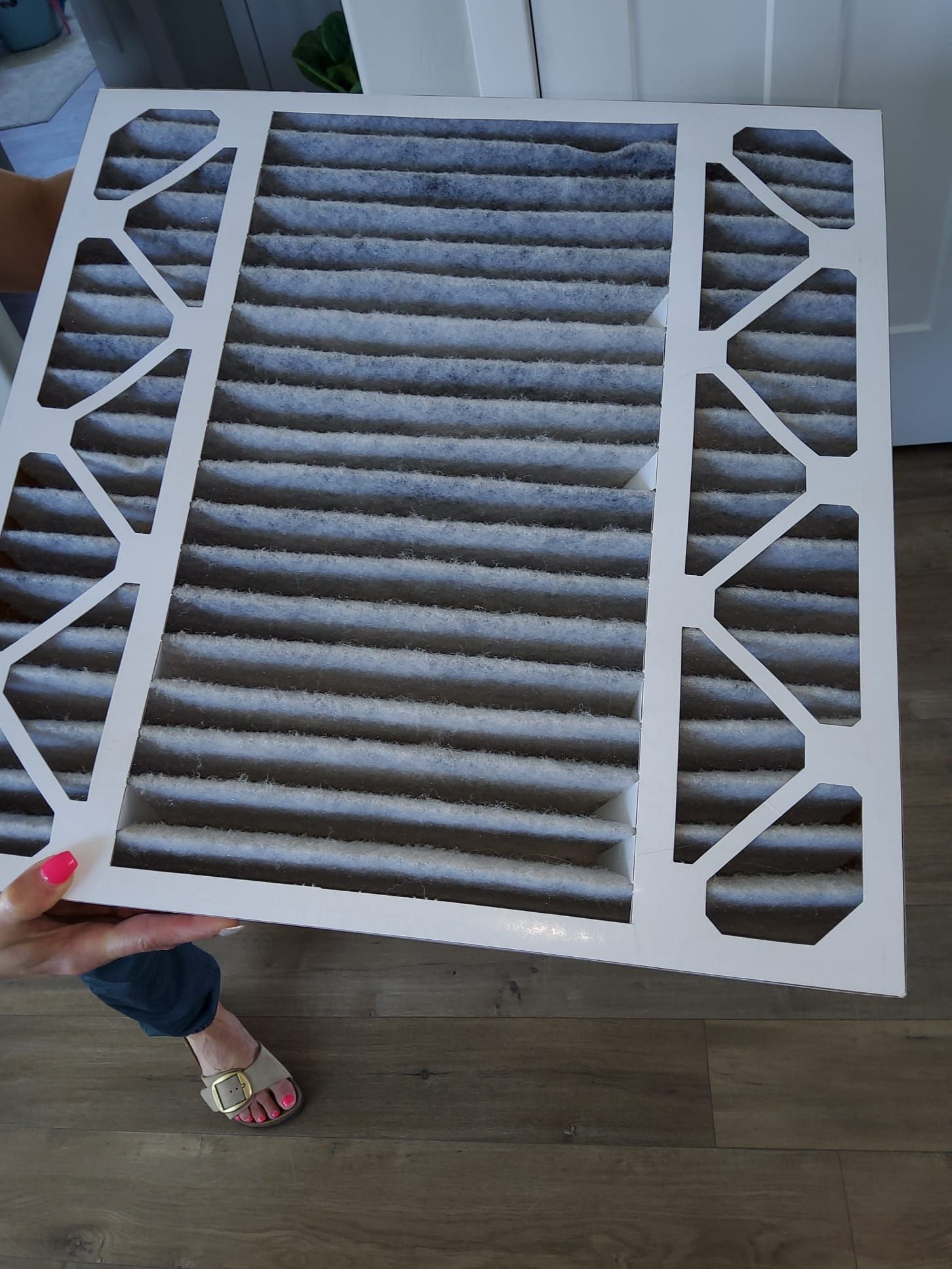 White HVAC air filter with gray pleated center, held above a wooden floor by a hand with red nail polish