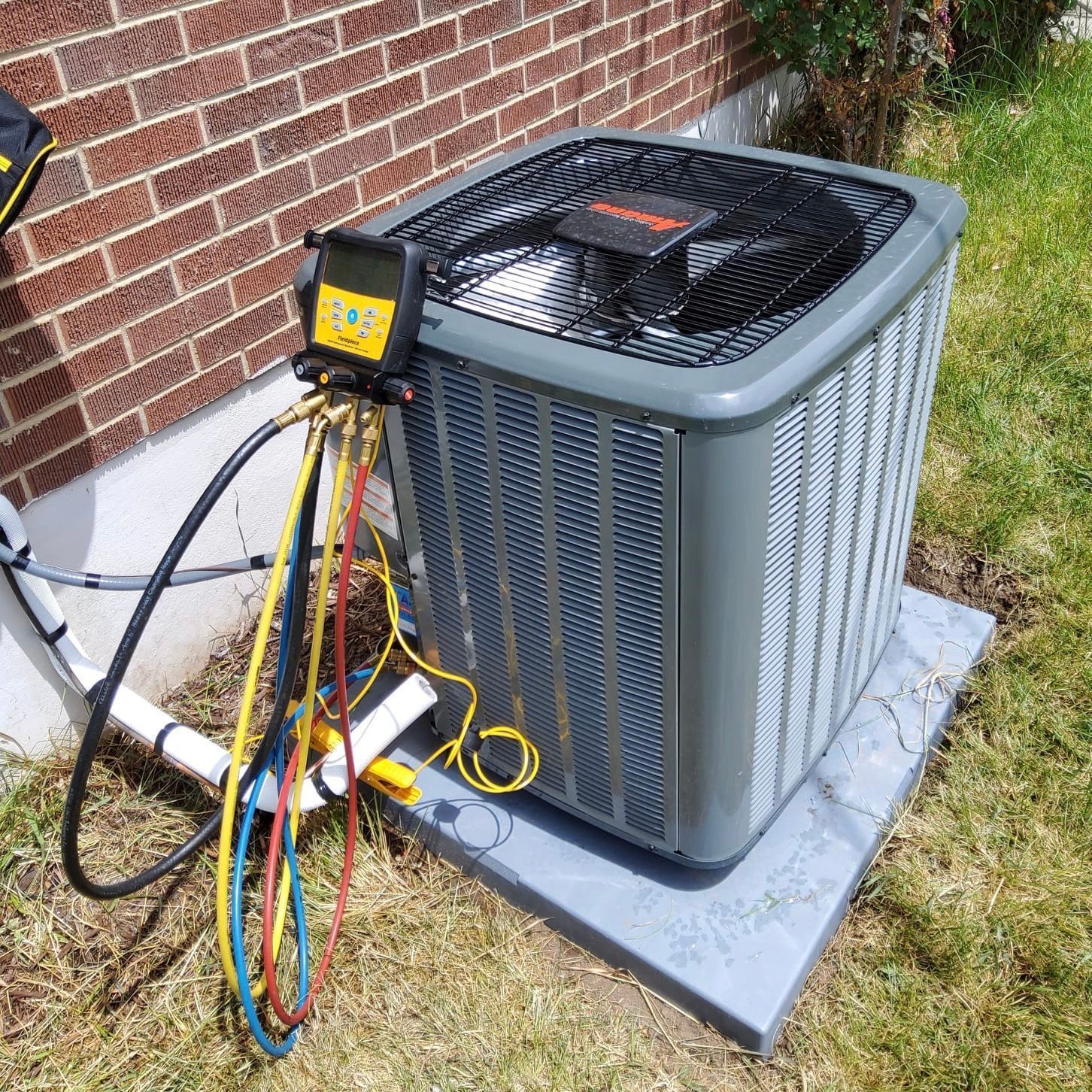 Outdoor HVAC unit with gauges and hoses attached, beside a brick wall and grass.