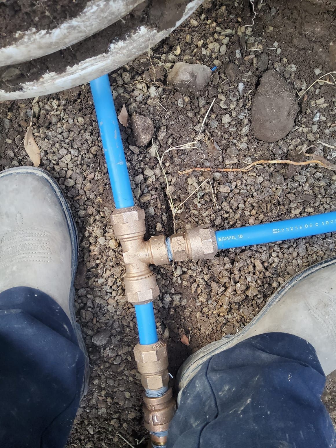 Blue PVC pipes and brass fittings forming a T-joint in dirt between two shoes.