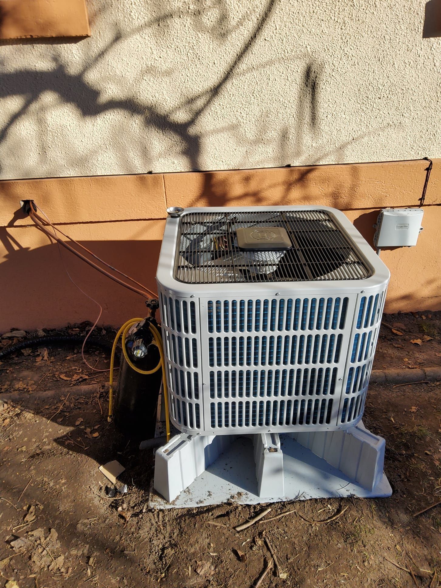 Outdoor HVAC condenser unit beside a tan wall, with a hose and shadowed branches nearby.