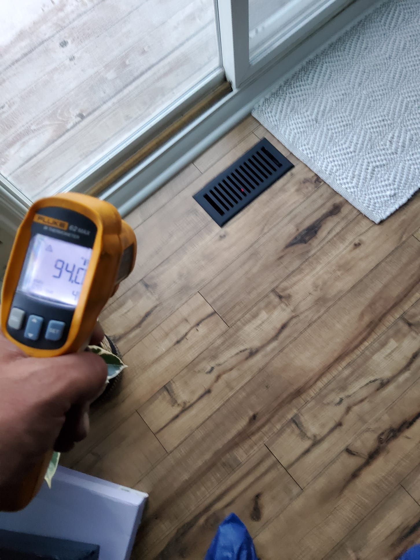 Handheld meter reading floor temperature near a sliding door and vent over wood flooring.