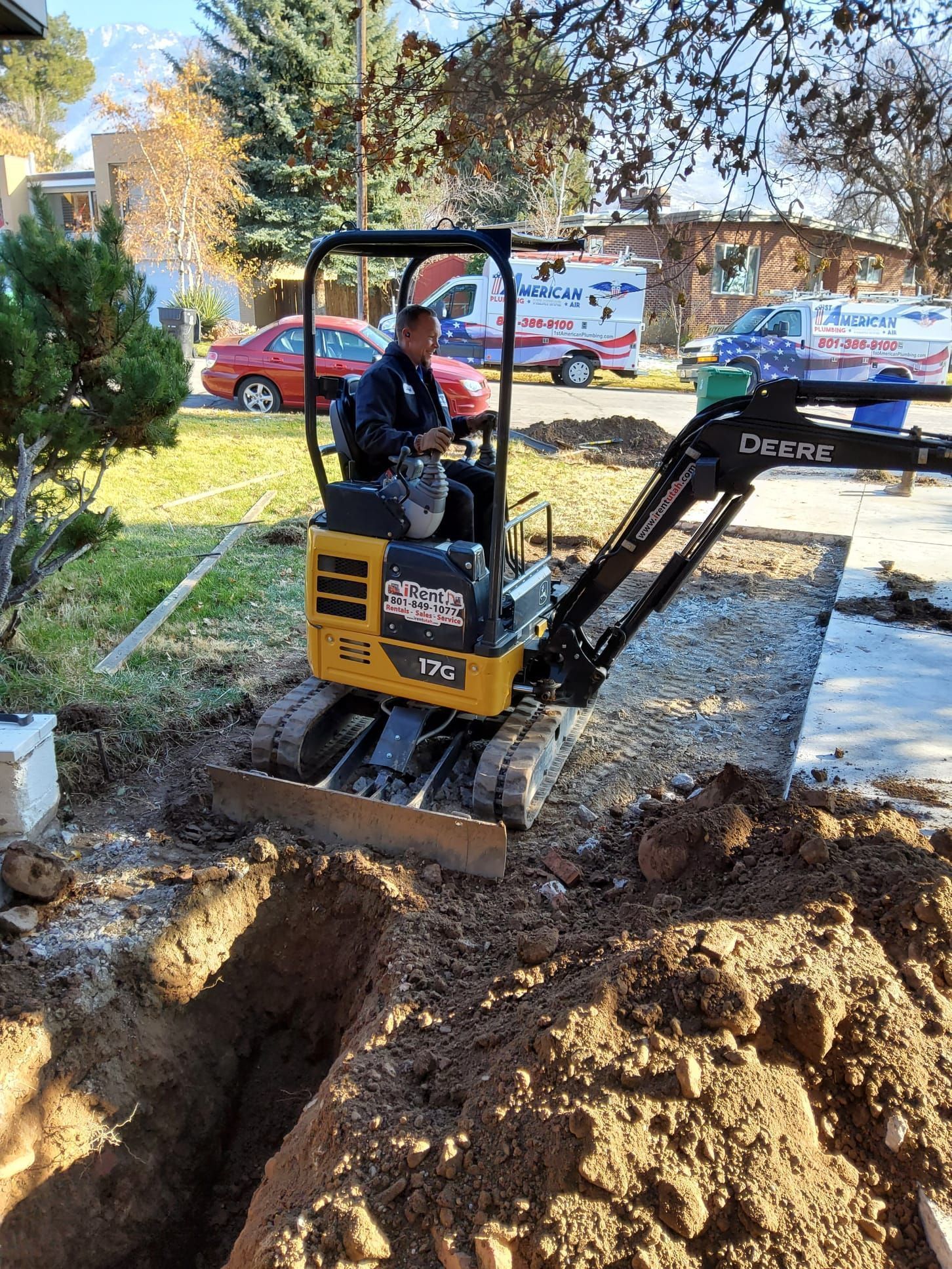 Mini excavator digging a muddy trench in a yard, with a worker operating it near a house.