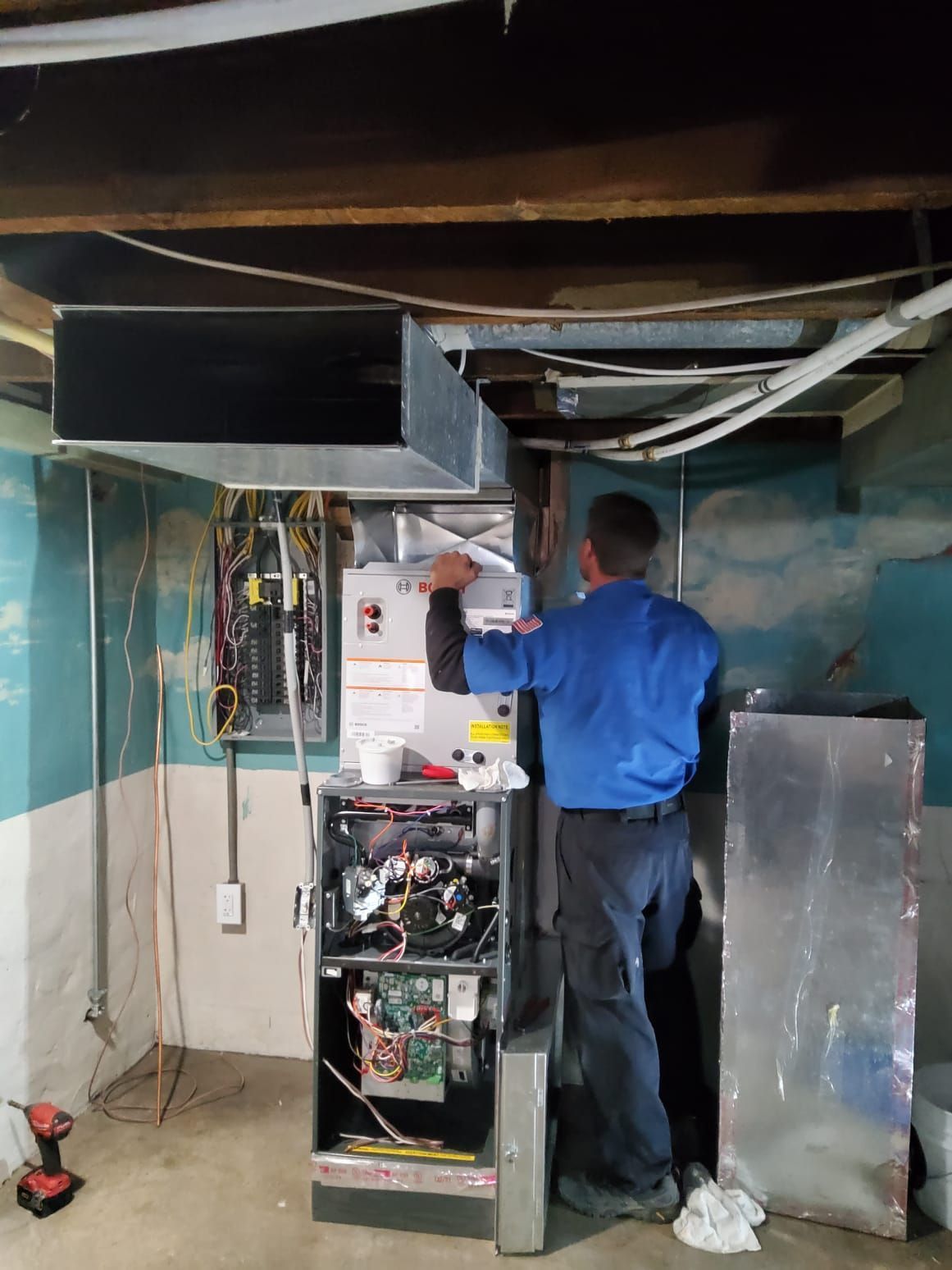 Technician working on electrical panels in a basement utility room with exposed wiring and ductwork