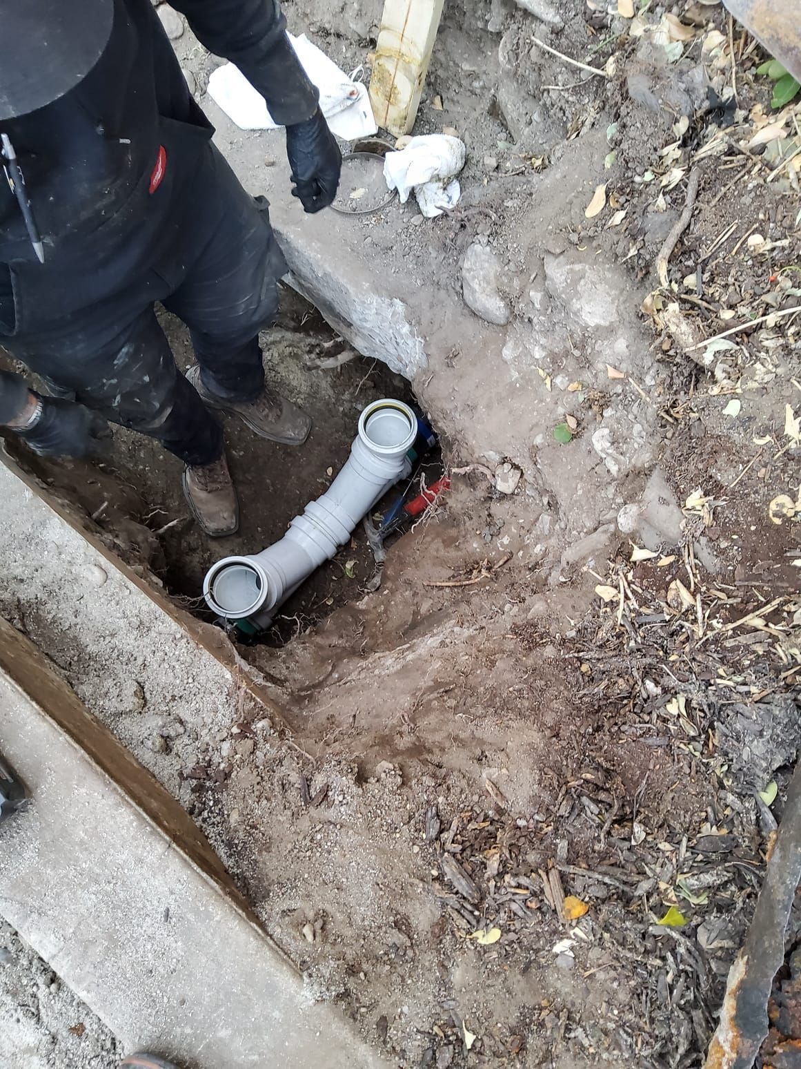 Worker beside exposed gray PVC pipe in muddy trench with debris and dirt walls
