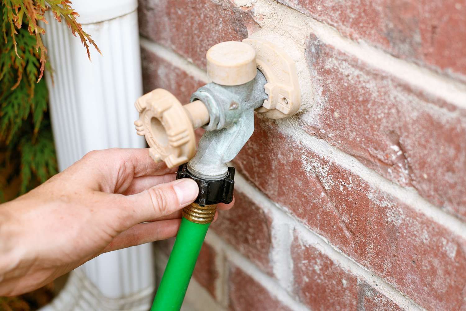 Hand turning on an outdoor faucet attached to a brick wall, with a green garden hose connected