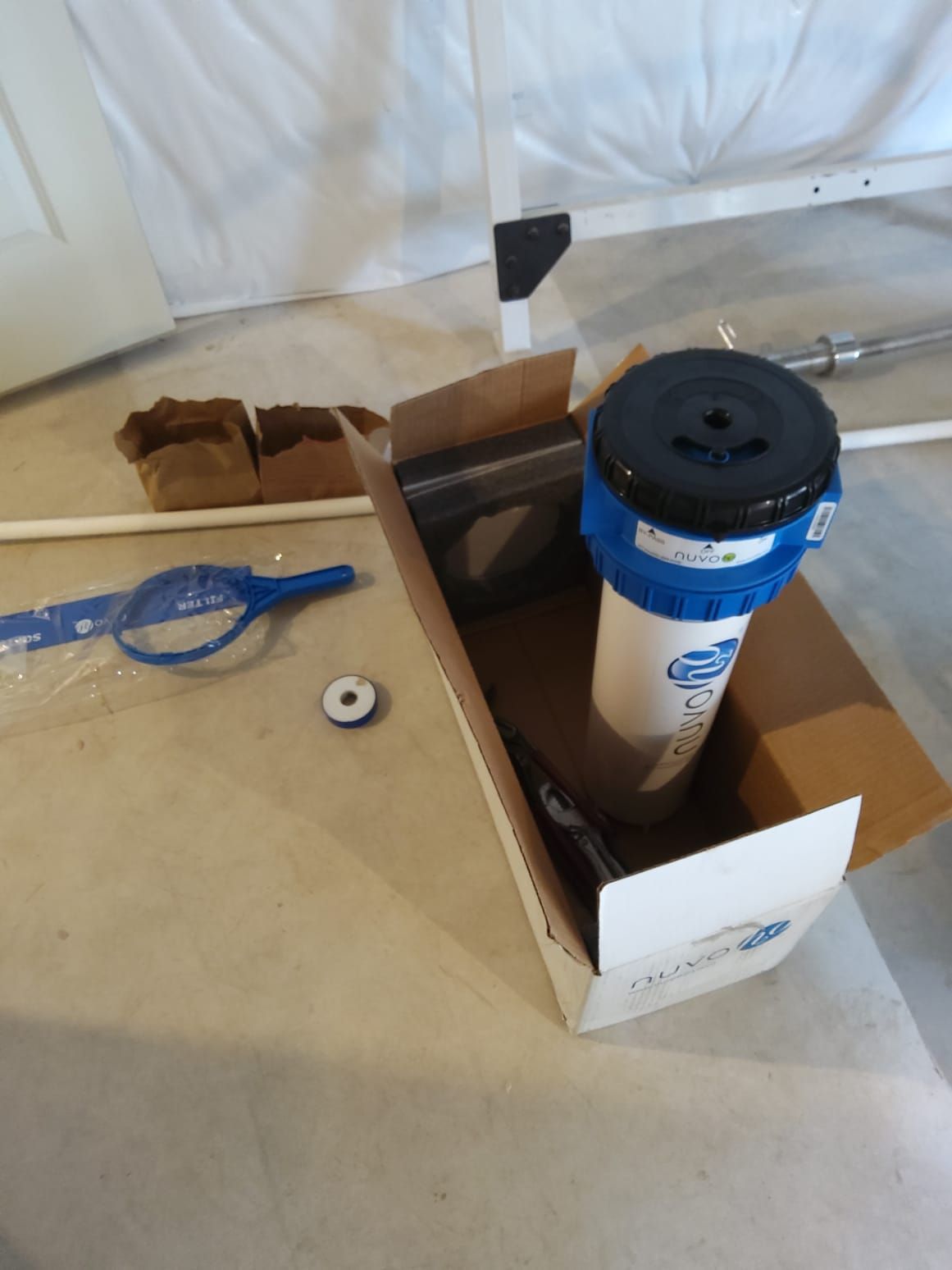 Open cardboard box holding a blue-and-white cylinder beside blue tool and small parts on a floor