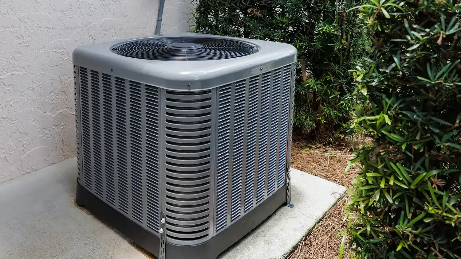 Outdoor air conditioner unit beside a stucco wall and shrubbery on a concrete pad