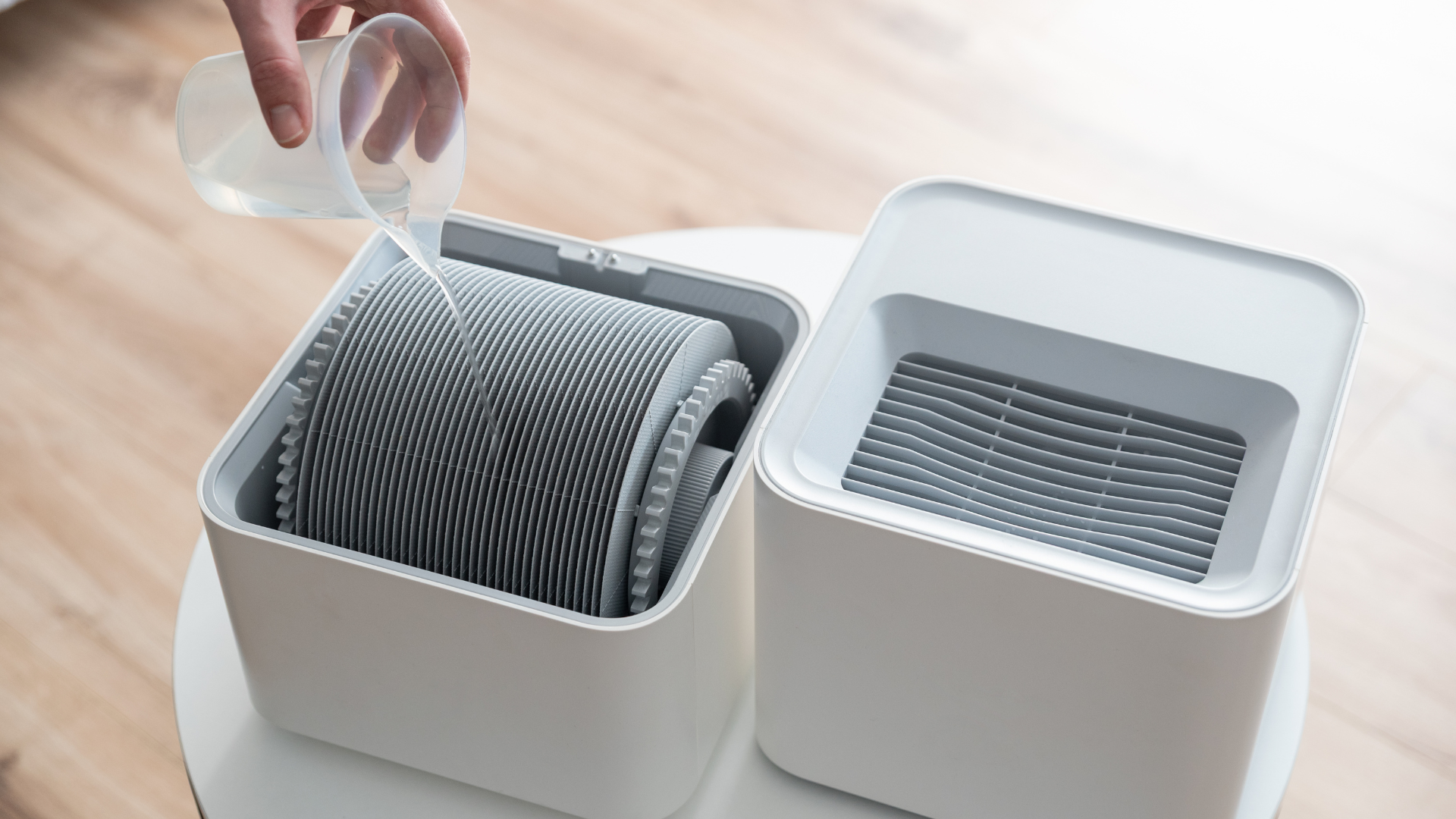 Hand placing a filter into a white air purifier on a wooden table, with another purifier beside it