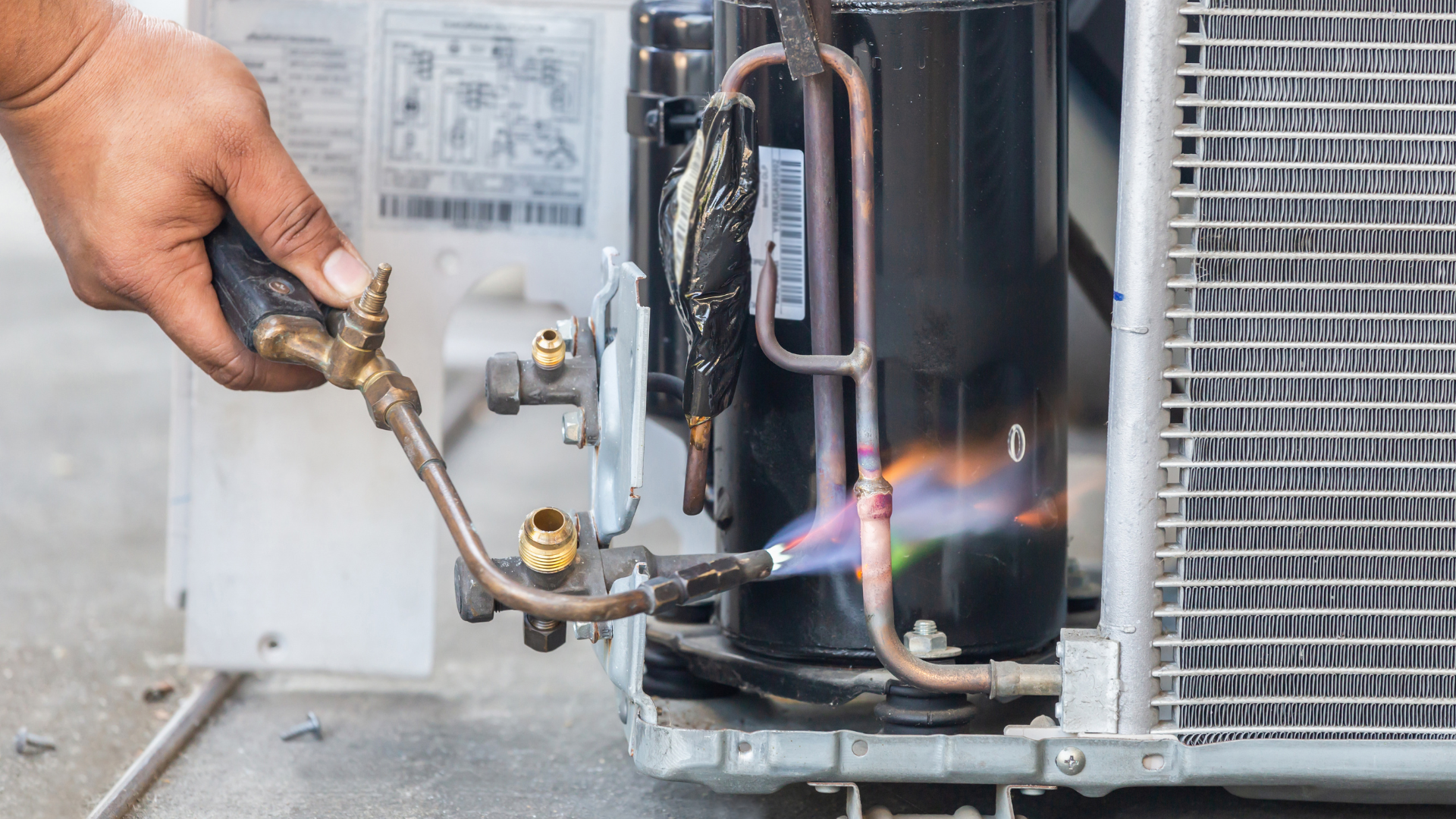 Hand torch heating a black metal pipe during HVAC repair, with a blue-orange flame near an outdoor unit
