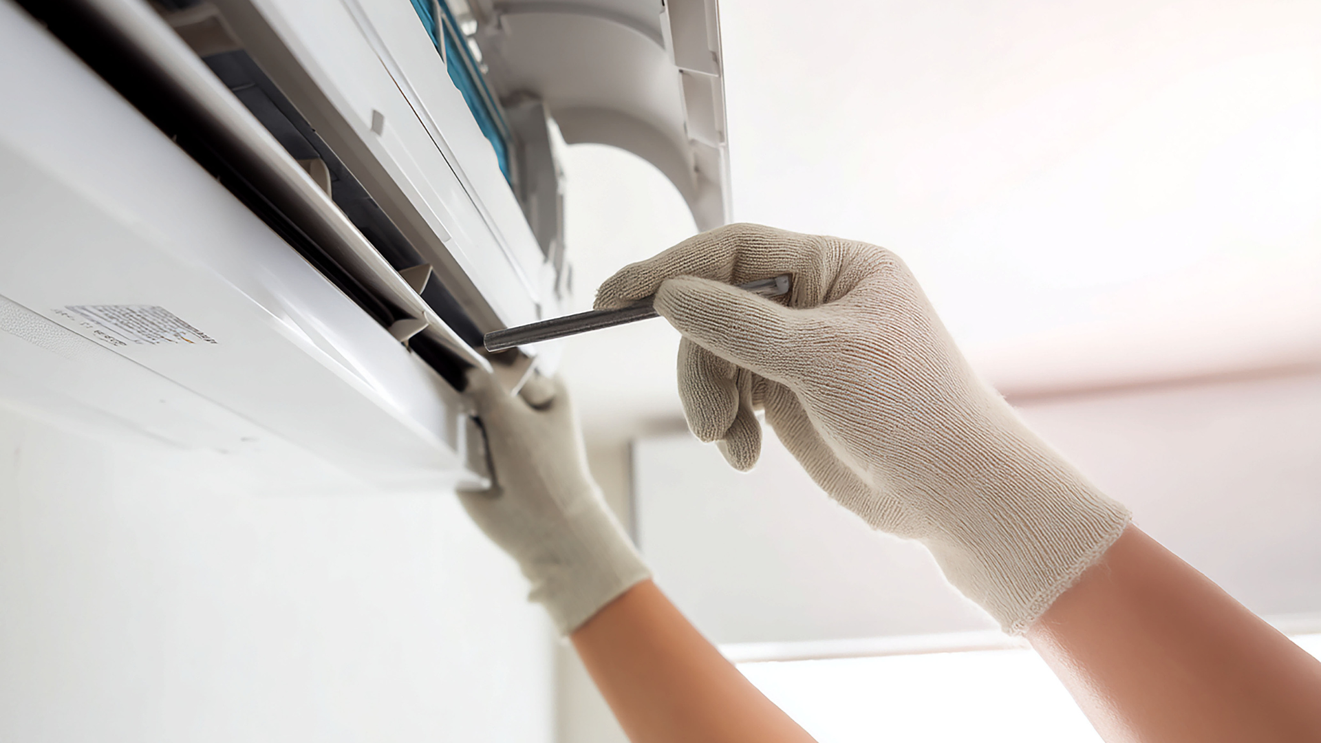 Gloved hands using a screwdriver to repair a white wall-mounted air conditioner unit