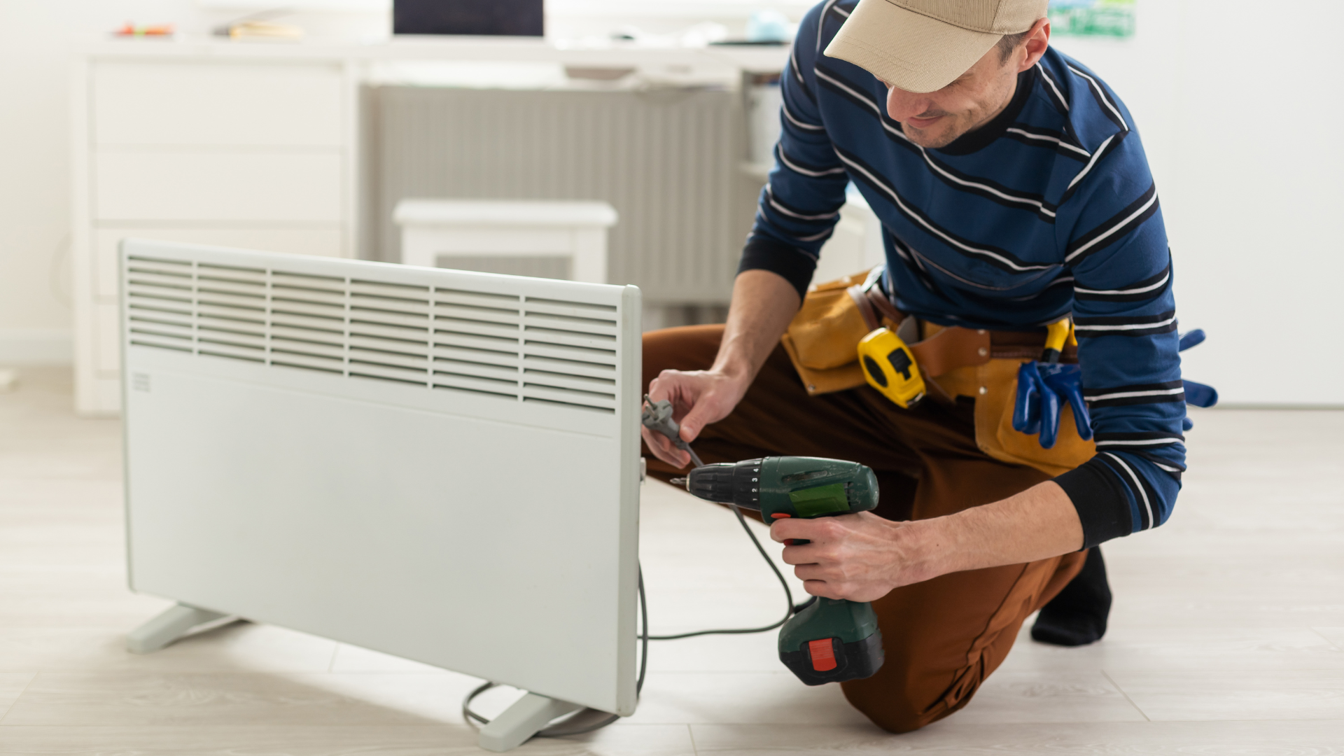 Technician kneeling beside a white radiator, using a drill to install or repair it indoors