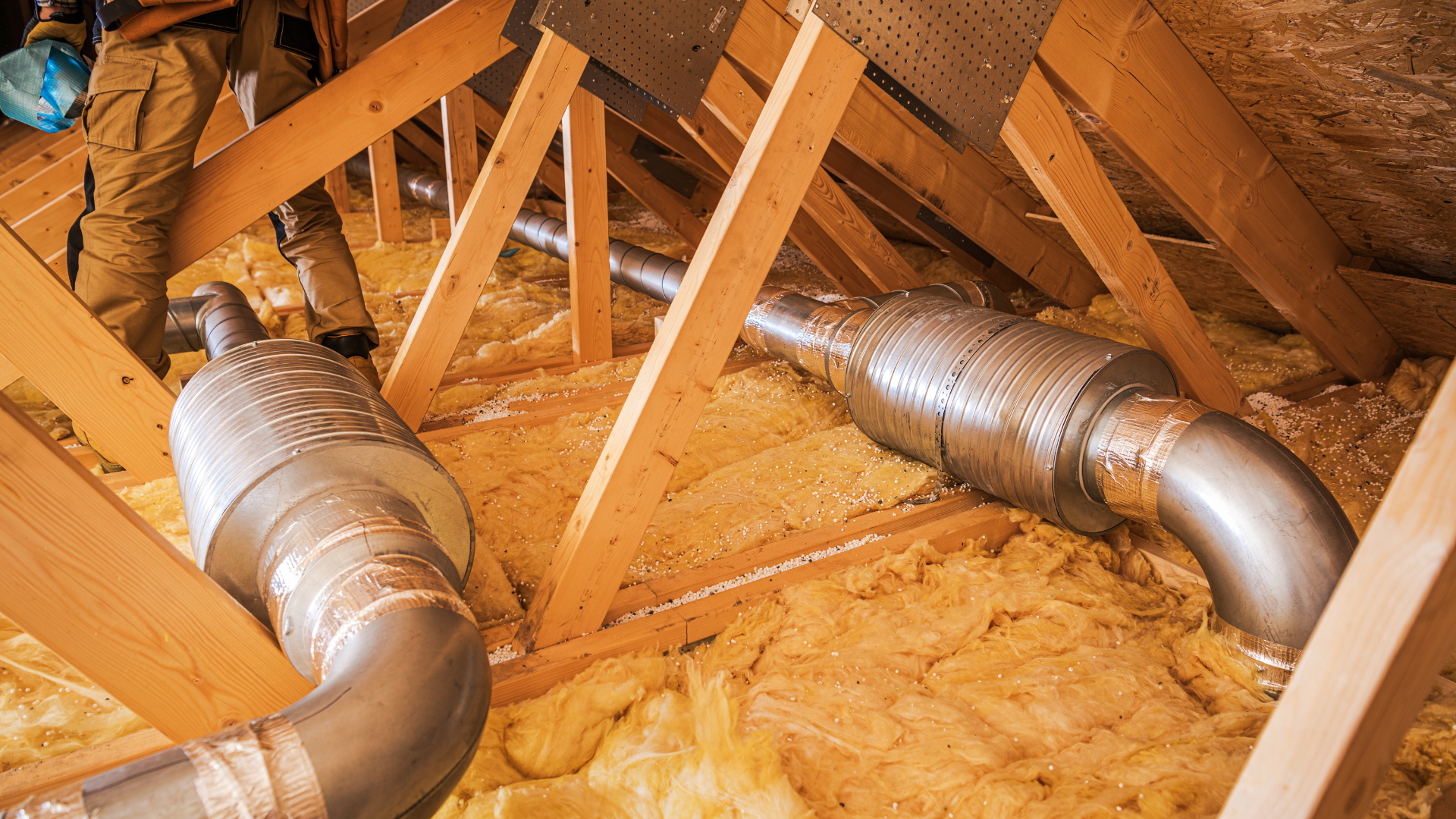 Insulated silver HVAC ducts running through an attic with exposed wooden rafters and yellow insulation.