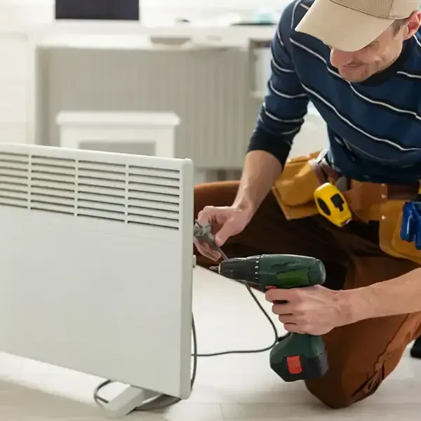 Technician kneeling beside a white radiator, using a drill to install or repair it indoors