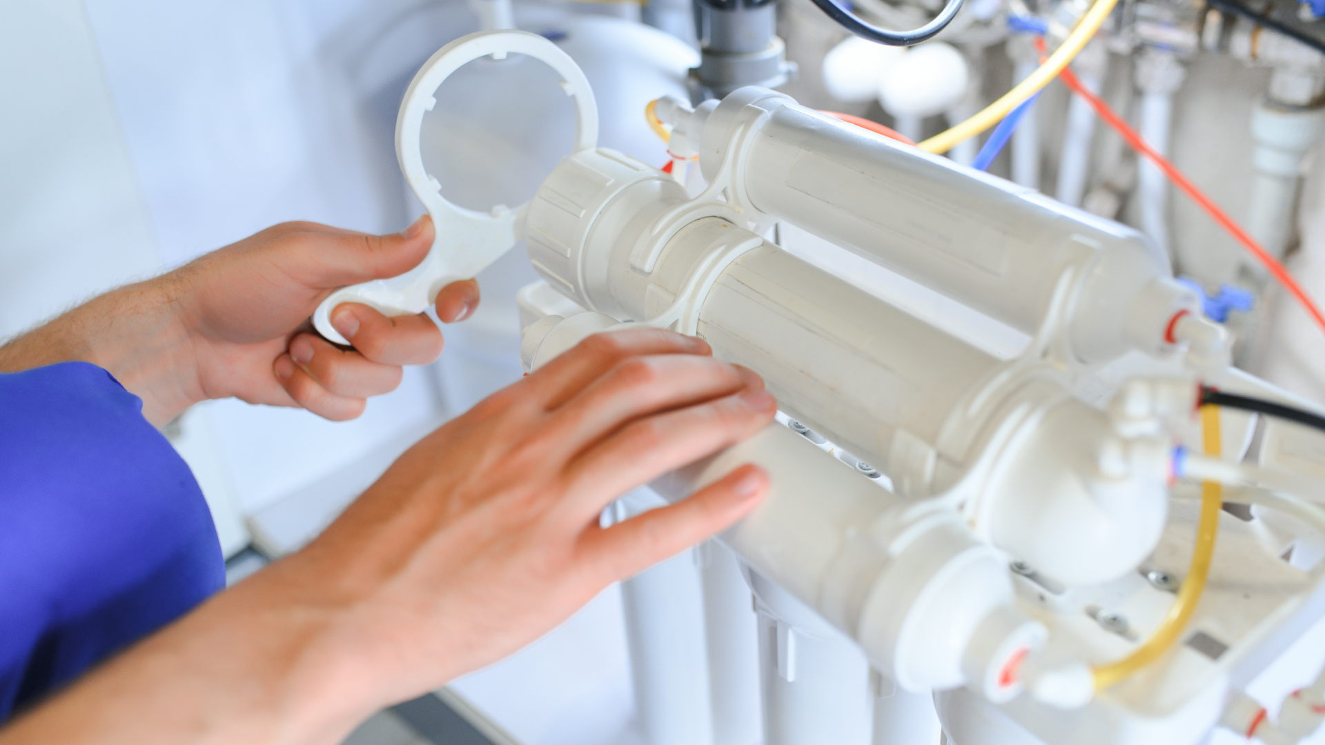 Hands adjusting a white industrial filter cartridge assembly with pipes and colored tubes in a lab setting