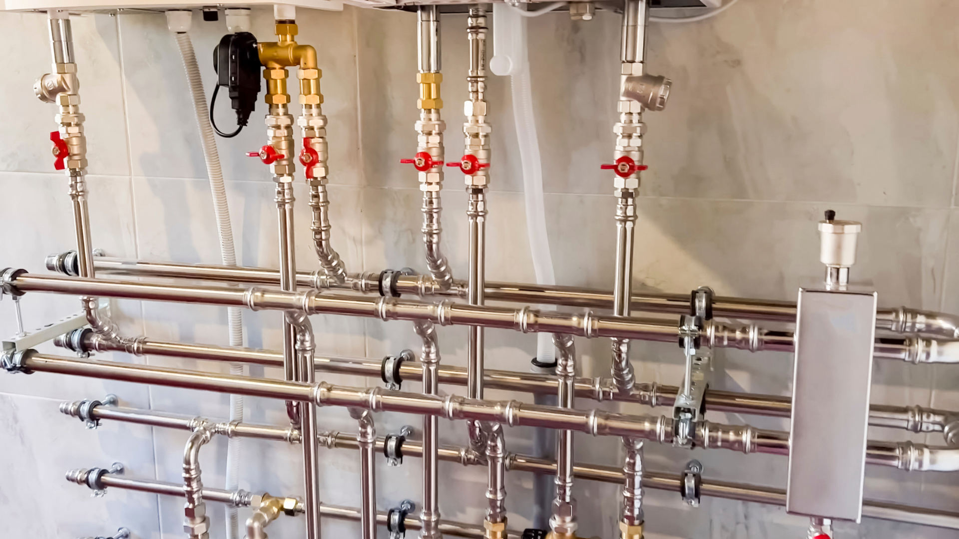 Metal pipes and valves on a wall in a utility room, with a white control box on the right.