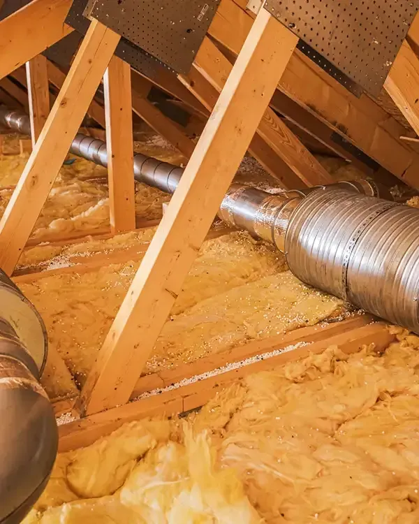 Insulated silver HVAC ducts running through an attic with exposed wooden rafters and yellow insulation.