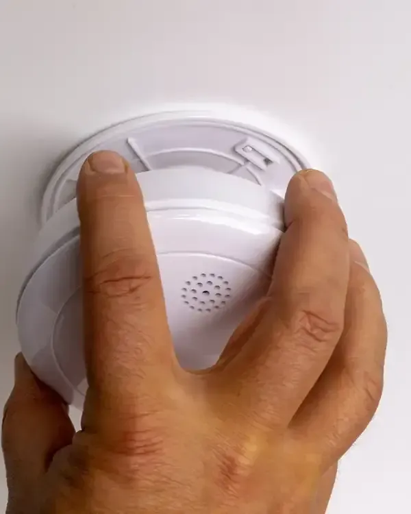 Hand installing a white smoke detector on a ceiling.